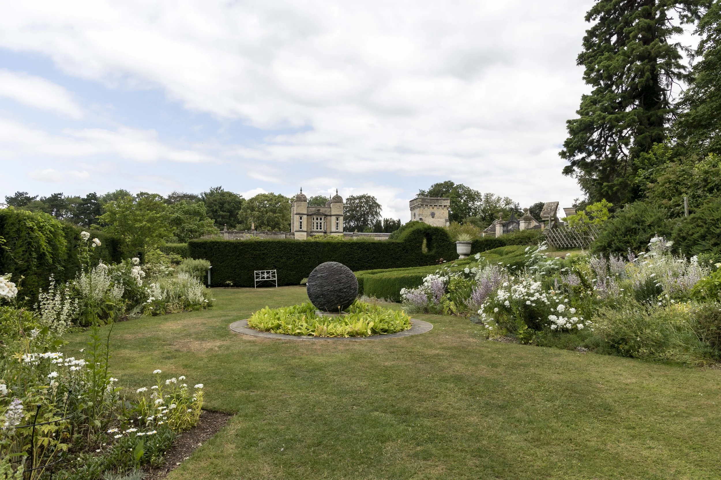A well-maintained garden with a round black sculpture on a circular base surrounded by lush greenery, trees, and historic buildings in the background under a partly cloudy sky.