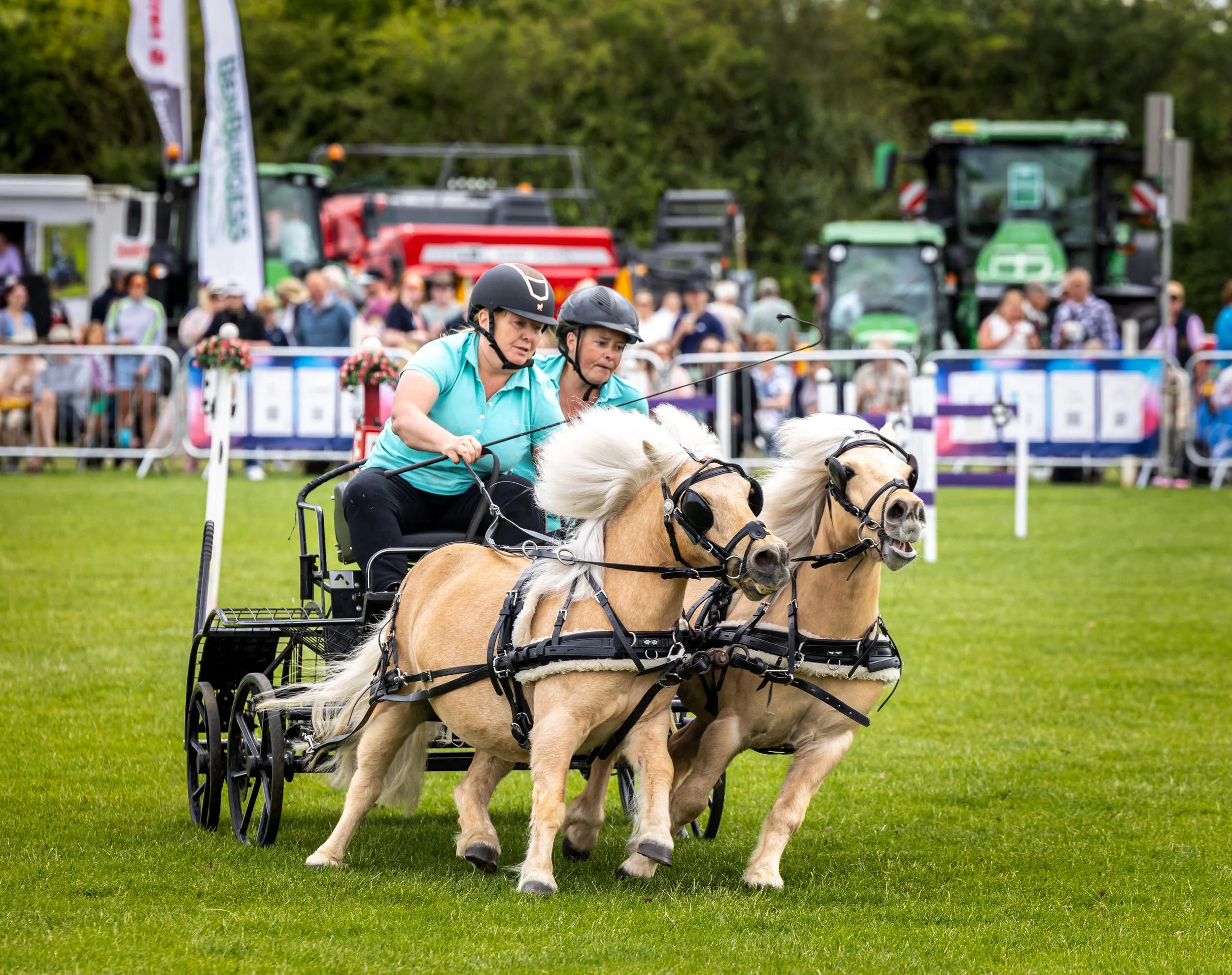 Two women riding a horse-drawn carriage at an outdoor event, with a crowd of spectators and parked tractors in the background.