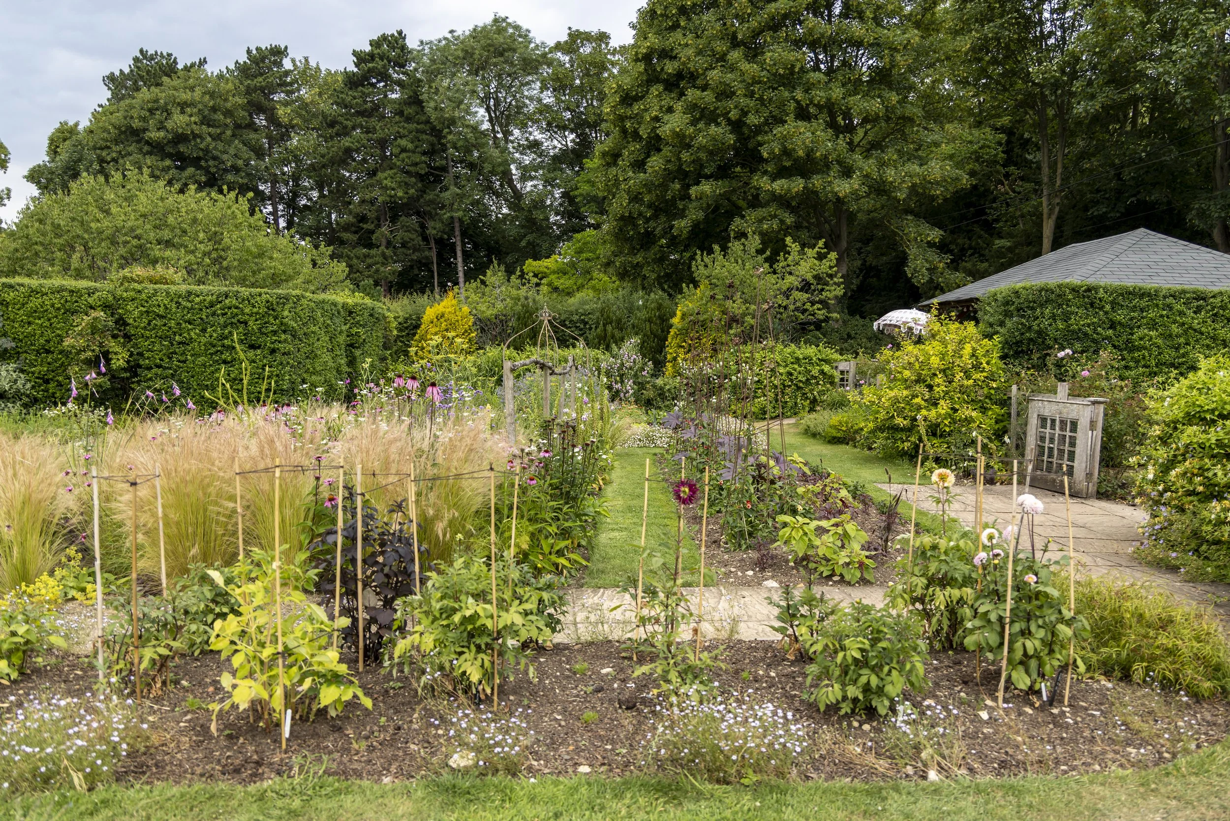 A lush garden with flowering plants, green bushes, and tall trees in the background. There is a stone pathway and a wooden fence surrounding the garden. A small gazebo with a gray roof is partially visible on the right side.