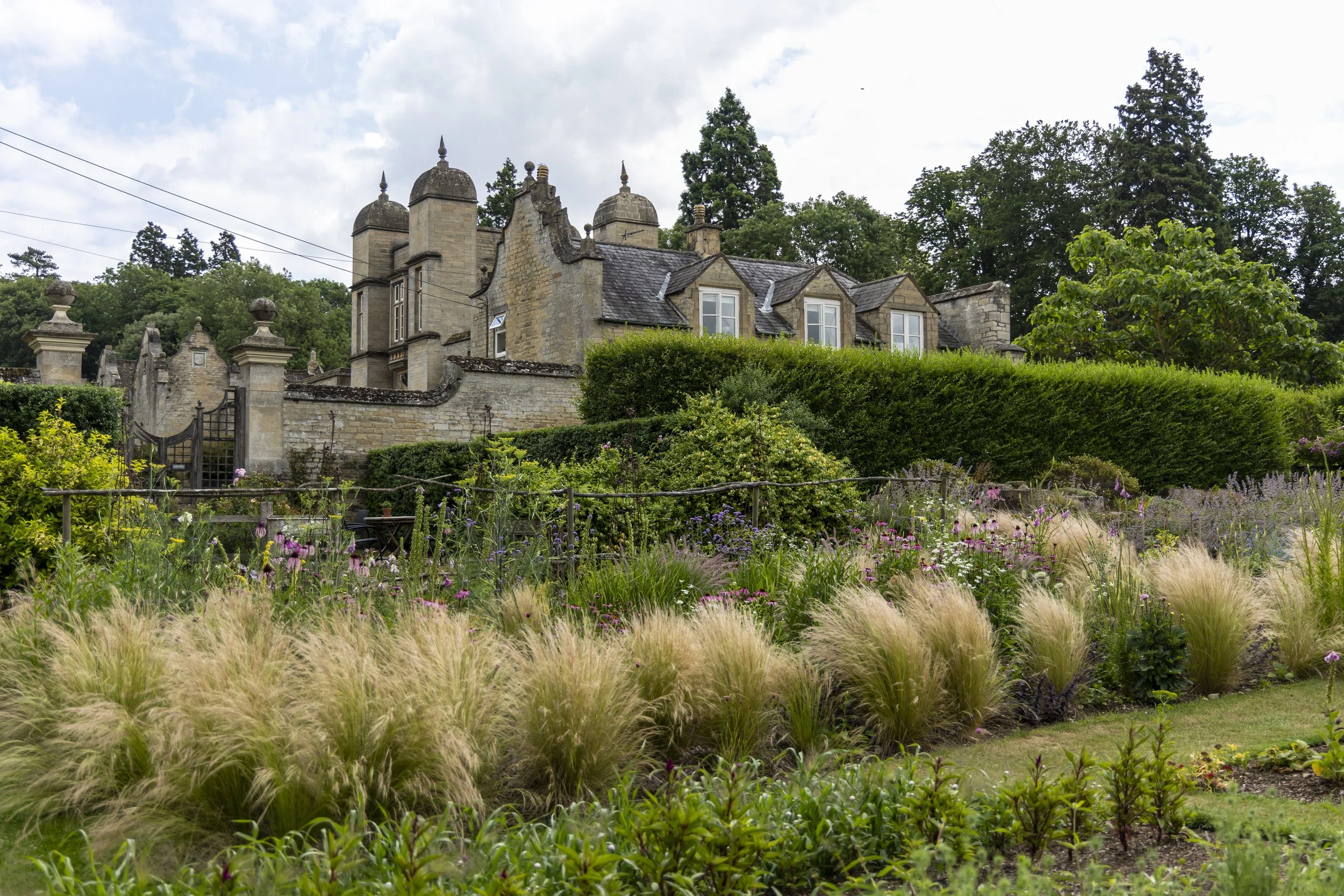 A historic stone mansion with multiple chimneys and turrets, surrounded by lush gardens and greenery, under a partly cloudy sky.