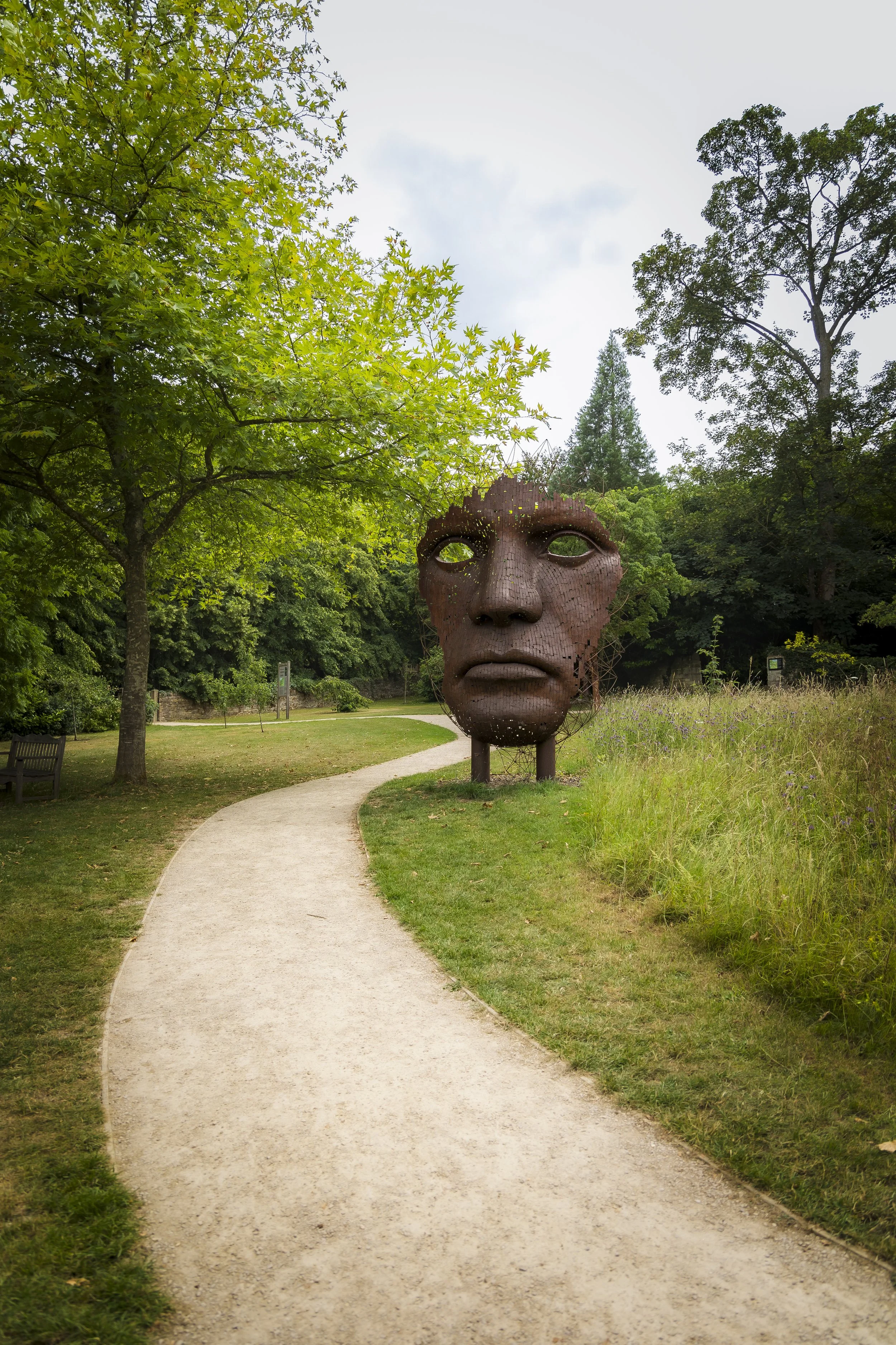 A large sculpture of a human face made of rusted metal stands in a park. The face has closed eyes and neutral expression. The park has a winding gravel path, green grass, trees, and bushes surrounding the sculpture under a cloudy sky at Burghley Hous