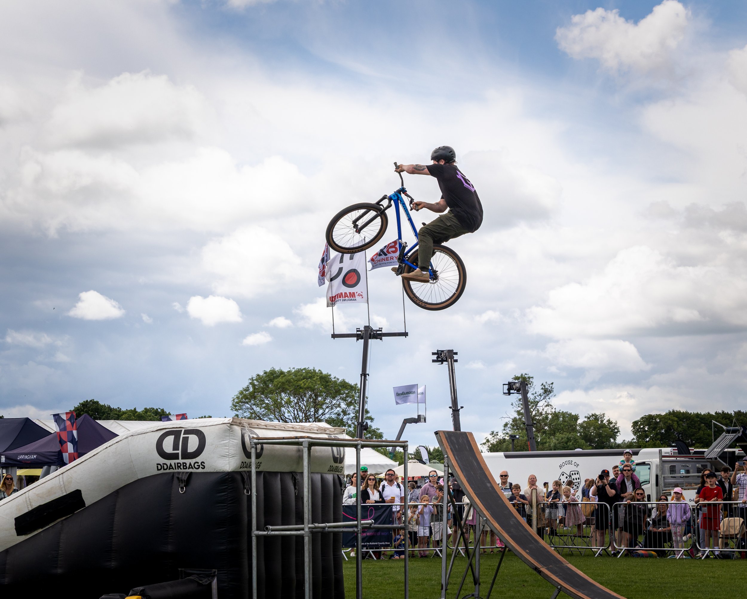 A man performing a bicycle stunt in the air at an outdoor event, with a crowd of spectators watching below. The sky is partly cloudy, and there are banners and tents in the background.