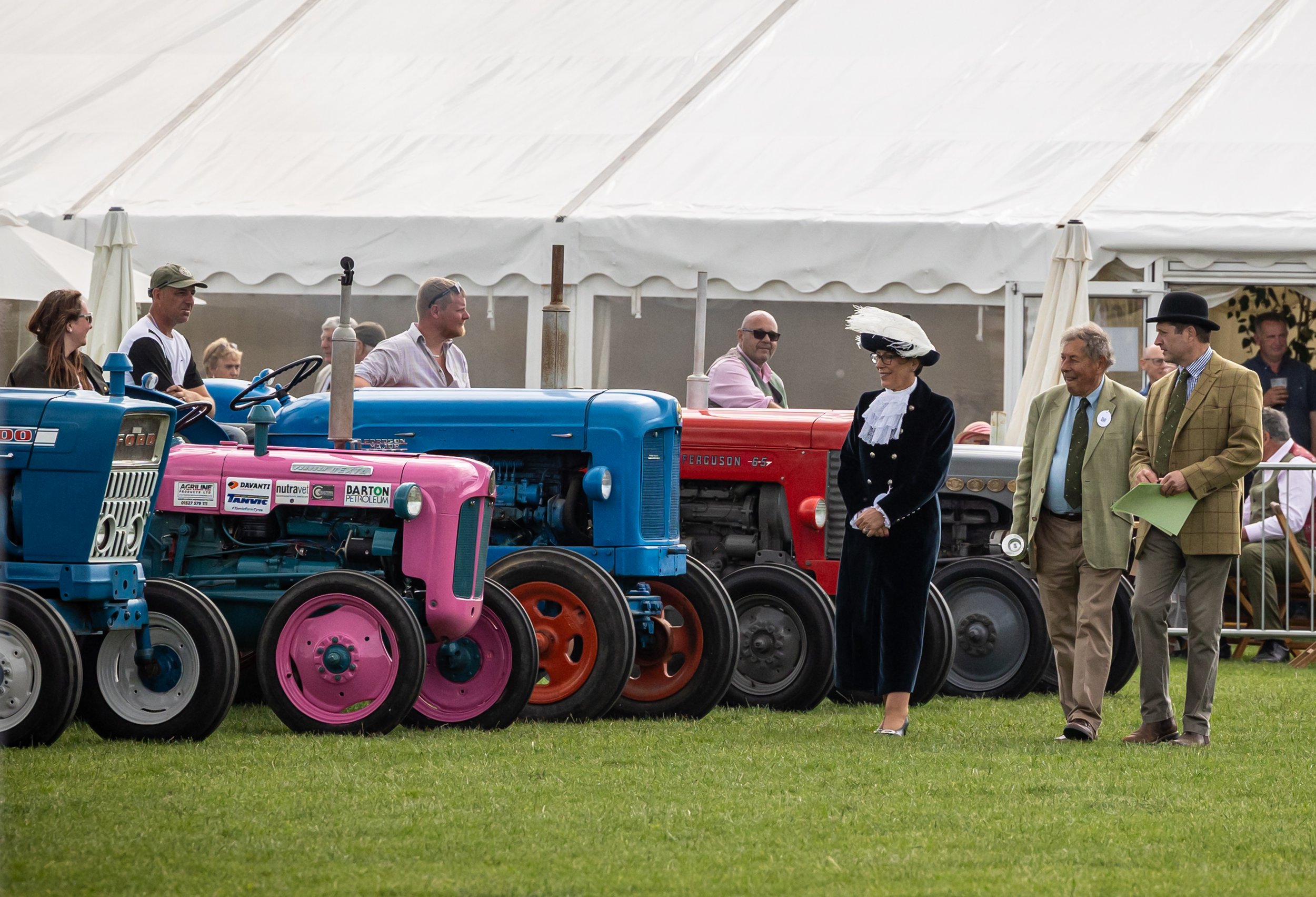 People at a vintage tractor show, including a woman in historical attire and men in formal clothing, with colorful tractors on display and a large white tent in the background.