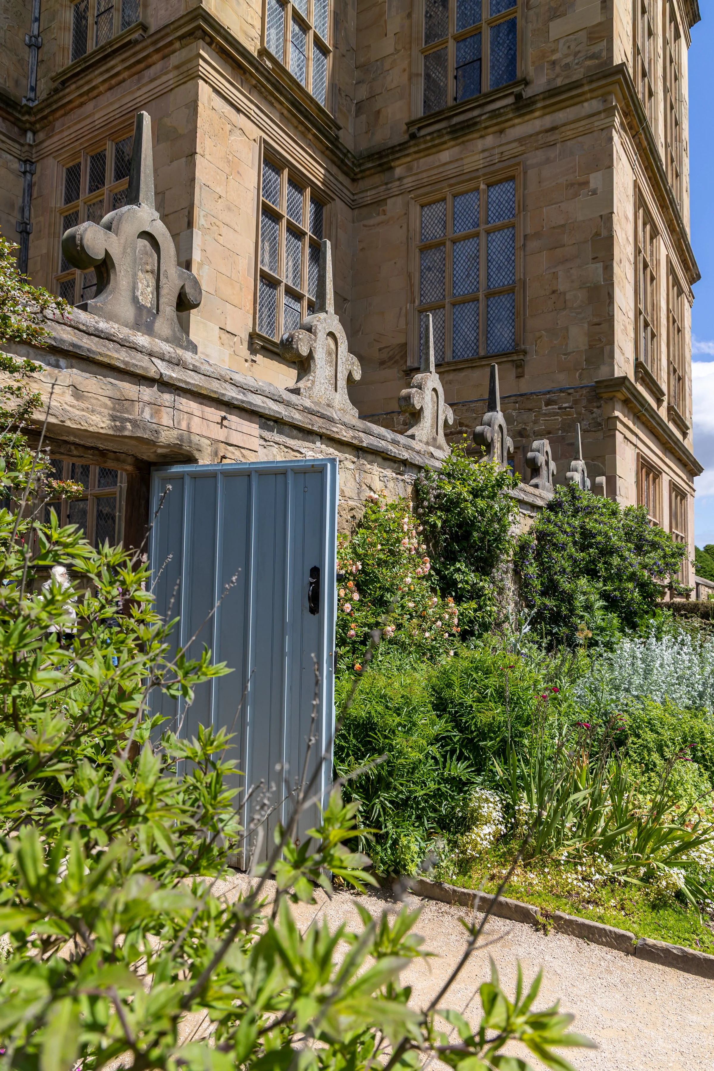 Hardwick Hall National Trust - View of a historic stone building with large leaded glass windows, stone decorative elements, and a garden area with lush plants and a partially open blue door.
