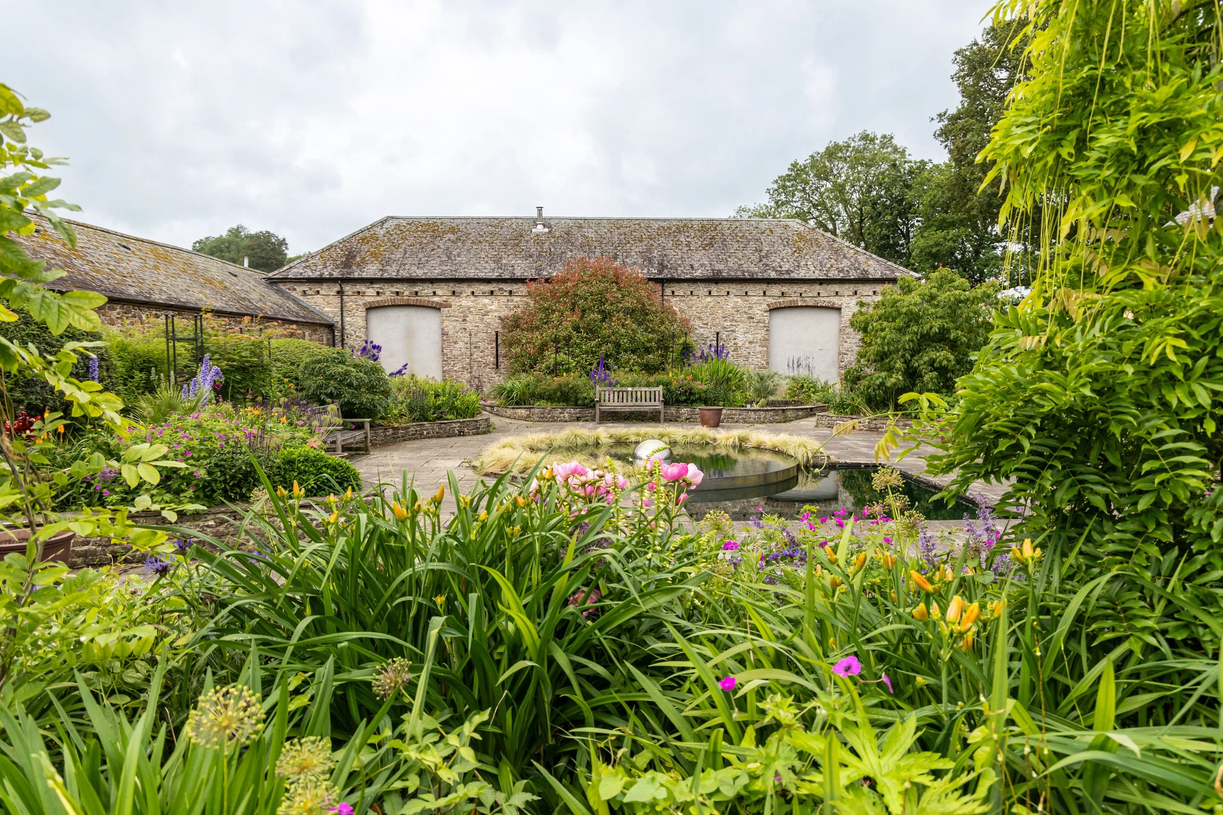 A garden with colorful flowers surrounding a pond, in front of a stone building with a sloped roof, behind trees and cloudy sky.