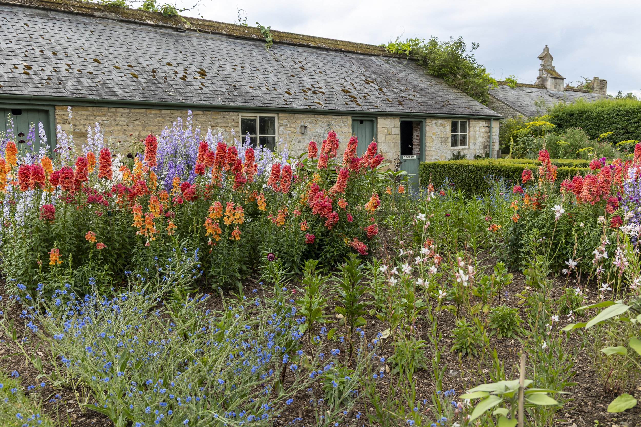 A rustic stone cottage with a slate roof, surrounded by colorful flowering plants and a neatly trimmed hedge under a cloudy sky.