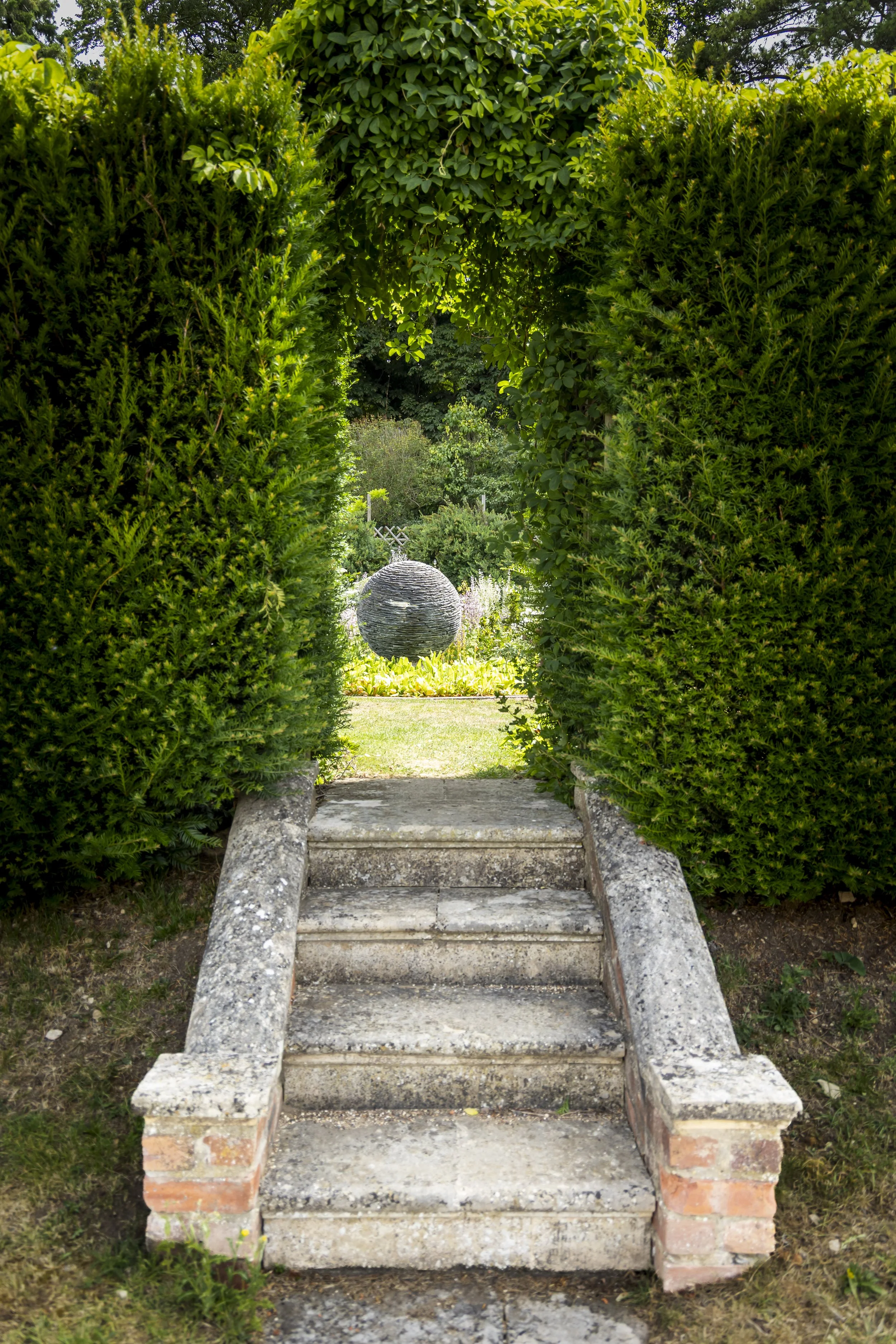 Concrete stairs with brick sides leading up to a garden with green bushes and trees. A round stone sculpture is visible in the background.