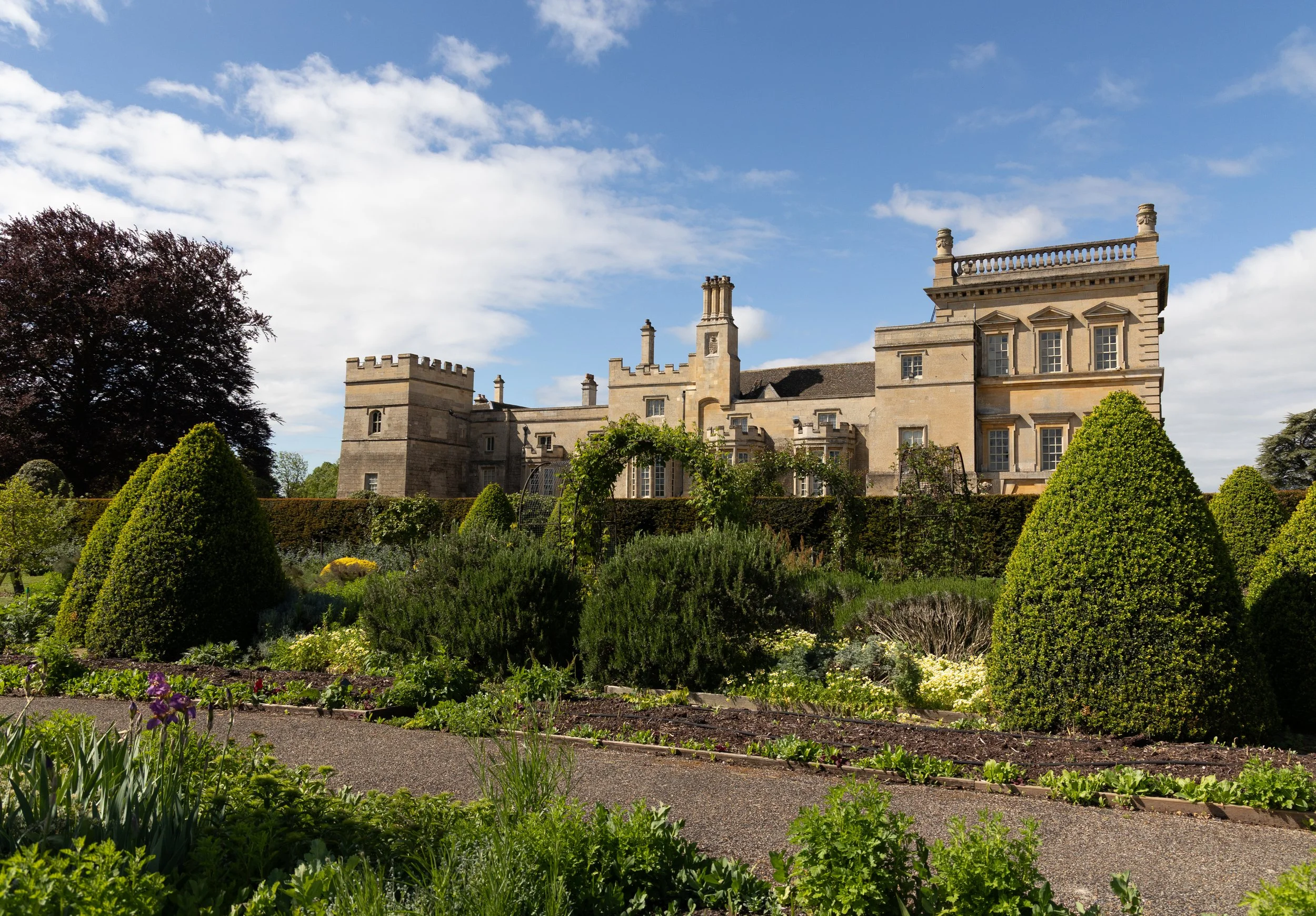 A historic castle with multiple towers and chimneys set among lush gardens with a gravel pathway and various green plants under a partly cloudy sky.