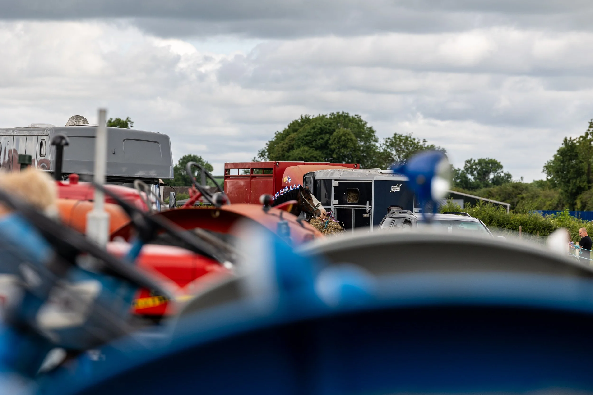 View of a parking area or outdoor event space with trailers, vehicles, and a horse with a decorative headband, under an overcast sky.