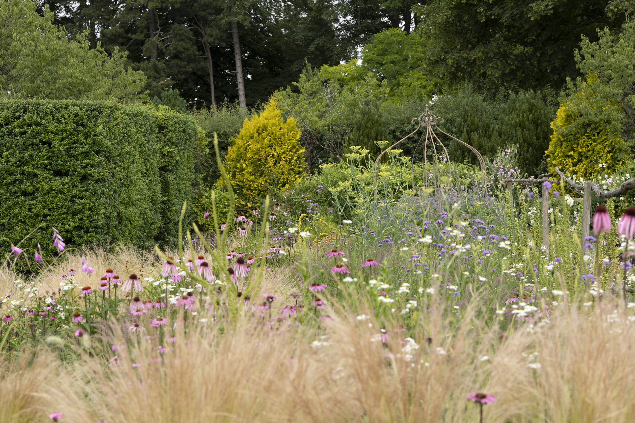 A garden with various colorful flowers in the foreground, green bushes and trees in the background, and a decorative metal sculpture among the plants.