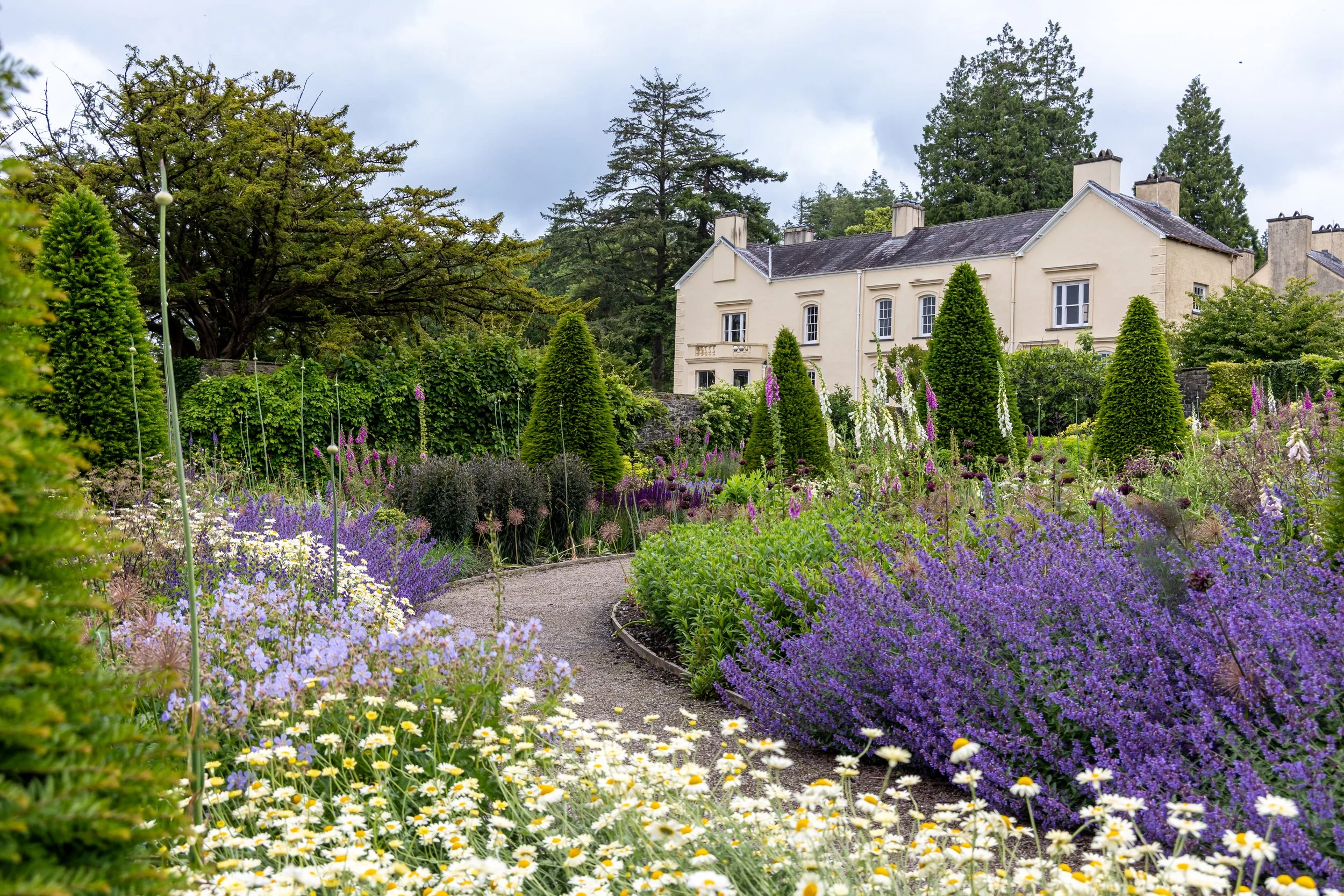 A large house surrounded by colorful flowering plants in a garden with many tall green trees background