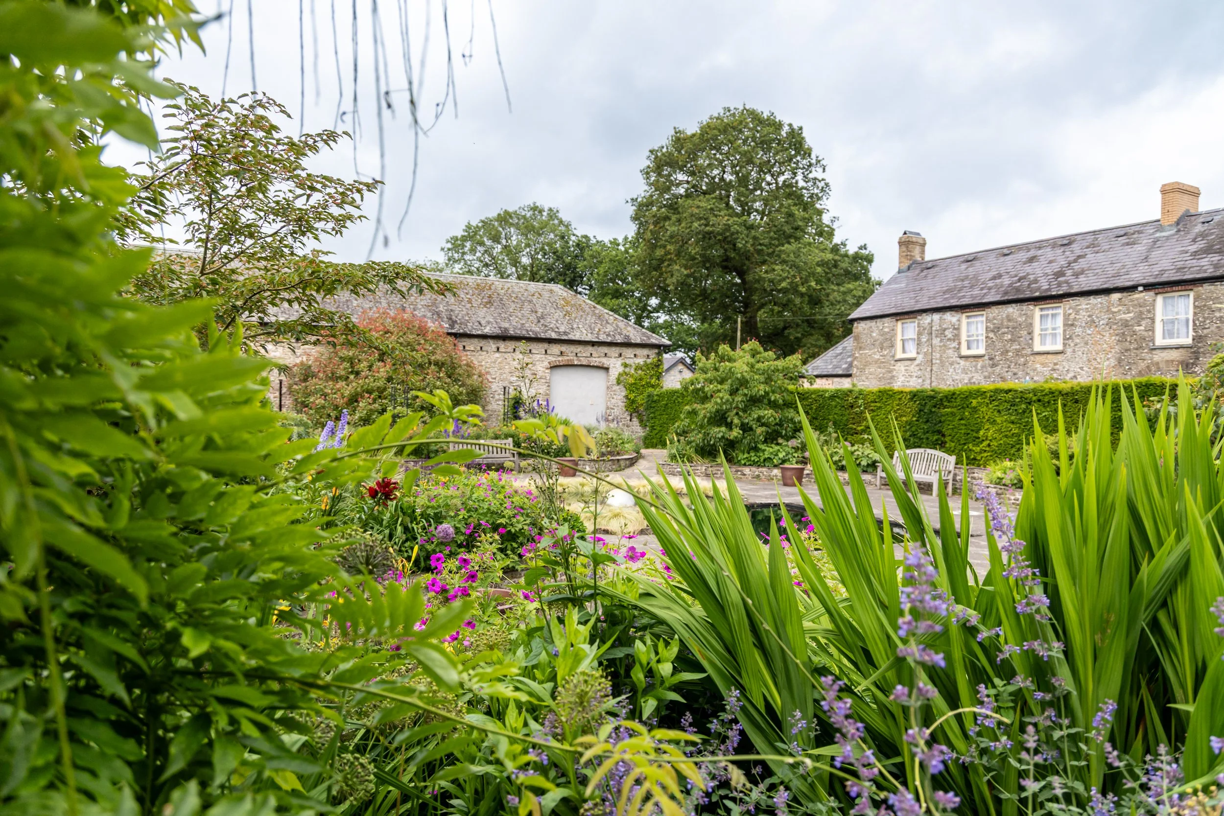 A scenic garden with lush green plants and colorful flowers in front of stone houses, large trees, and a cloudy sky.