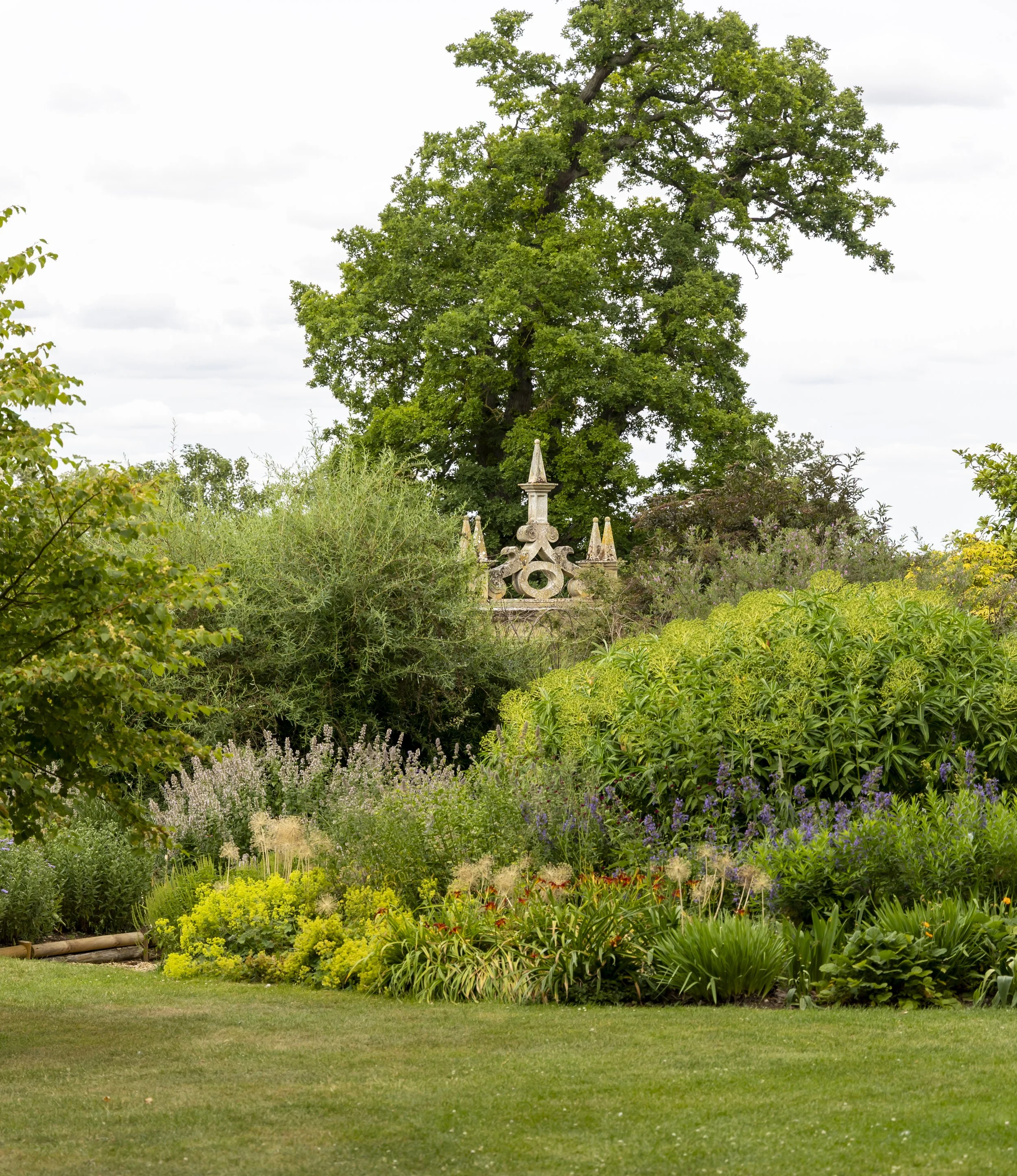 A garden with a variety of green plants and flowers, featuring a large tree in the background and decorative stone structures amid the foliage.