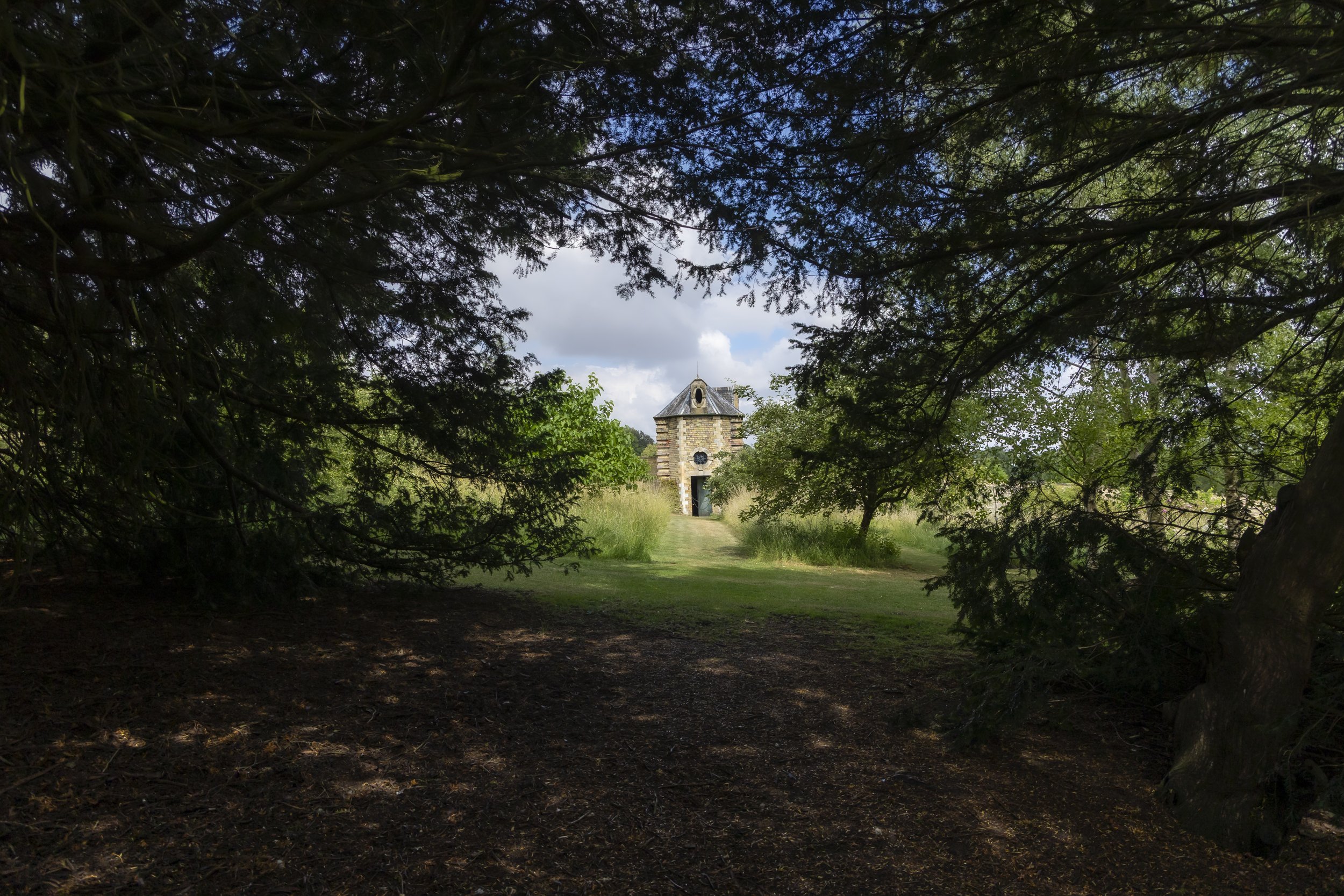 A small stone building with a pointed roof, surrounded by trees and greenery, viewed through a natural arch made of branches and leaves.
