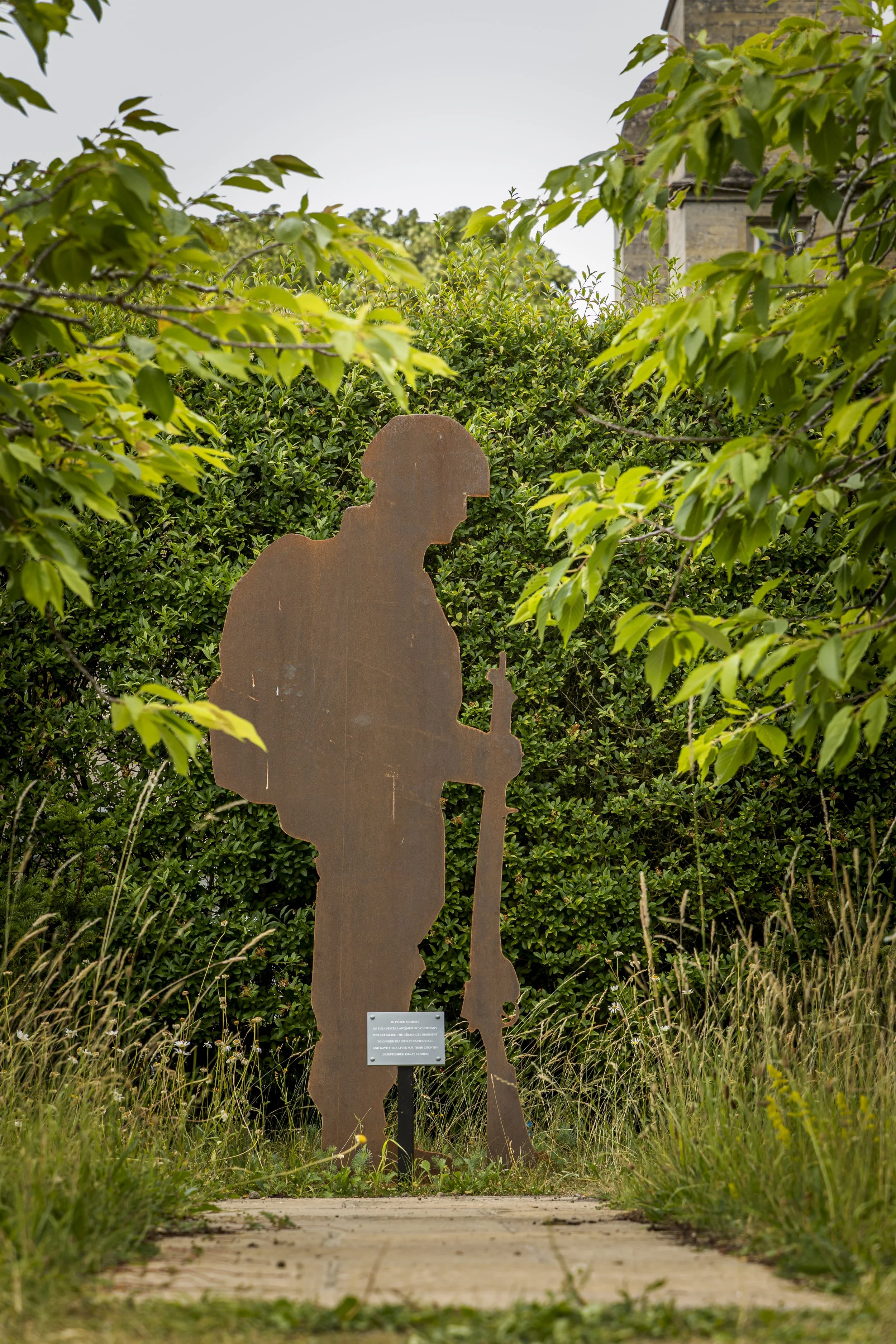 Steel silhouette sculpture of a soldier holding a rifle, placed in a garden with green bushes and trees, and a building in the background.