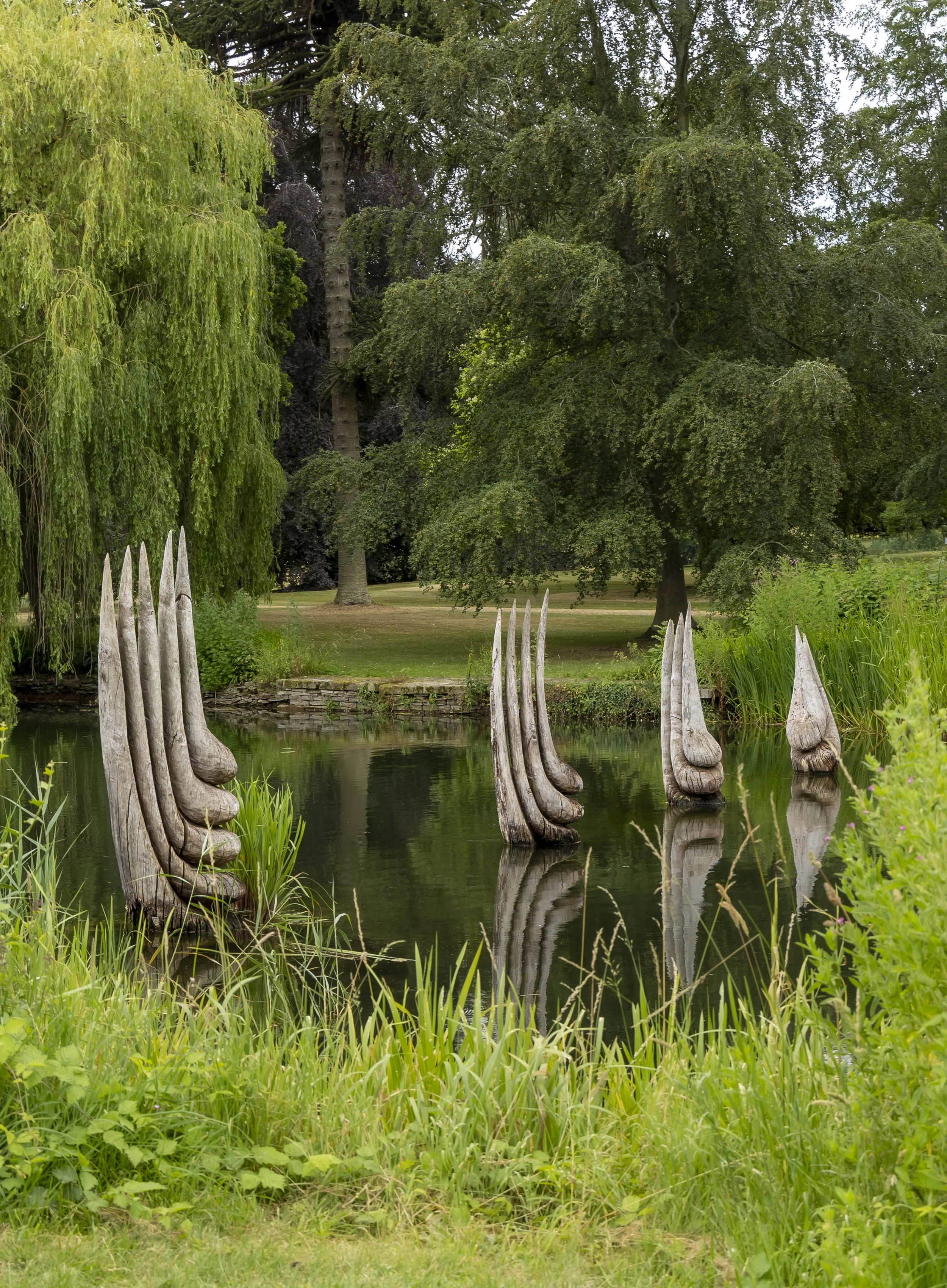A pond surrounded by green grass, trees, and plants. There are wooden posts emerging from the water, creating a mirror reflection of their shape. The scene is peaceful and lush with a variety of foliage.