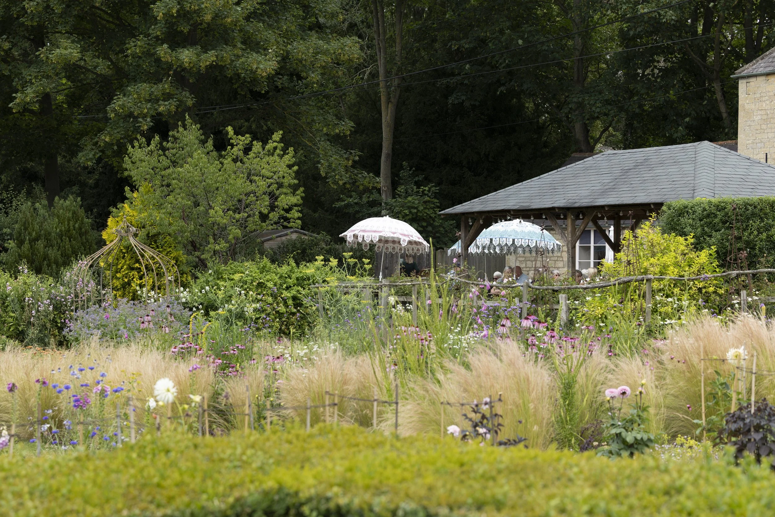 A lush garden scene with colorful flowers, a white decorative gazebo, and umbrellas for outdoor seating, surrounded by trees and greenery.