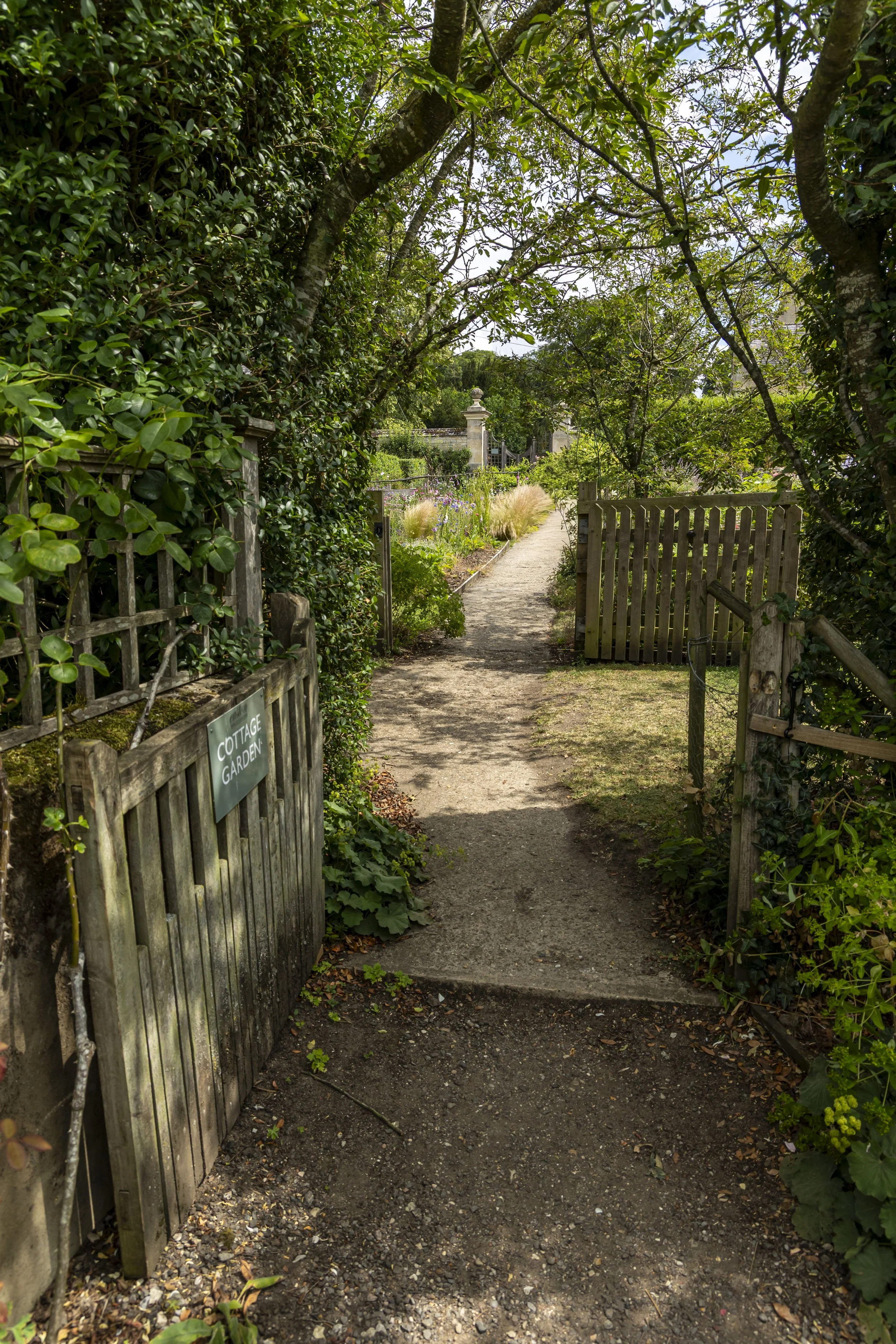 A garden pathway lined with green plants and trees, with a wooden gate on the right and a sign on the left that says 'Cottage Garden'.