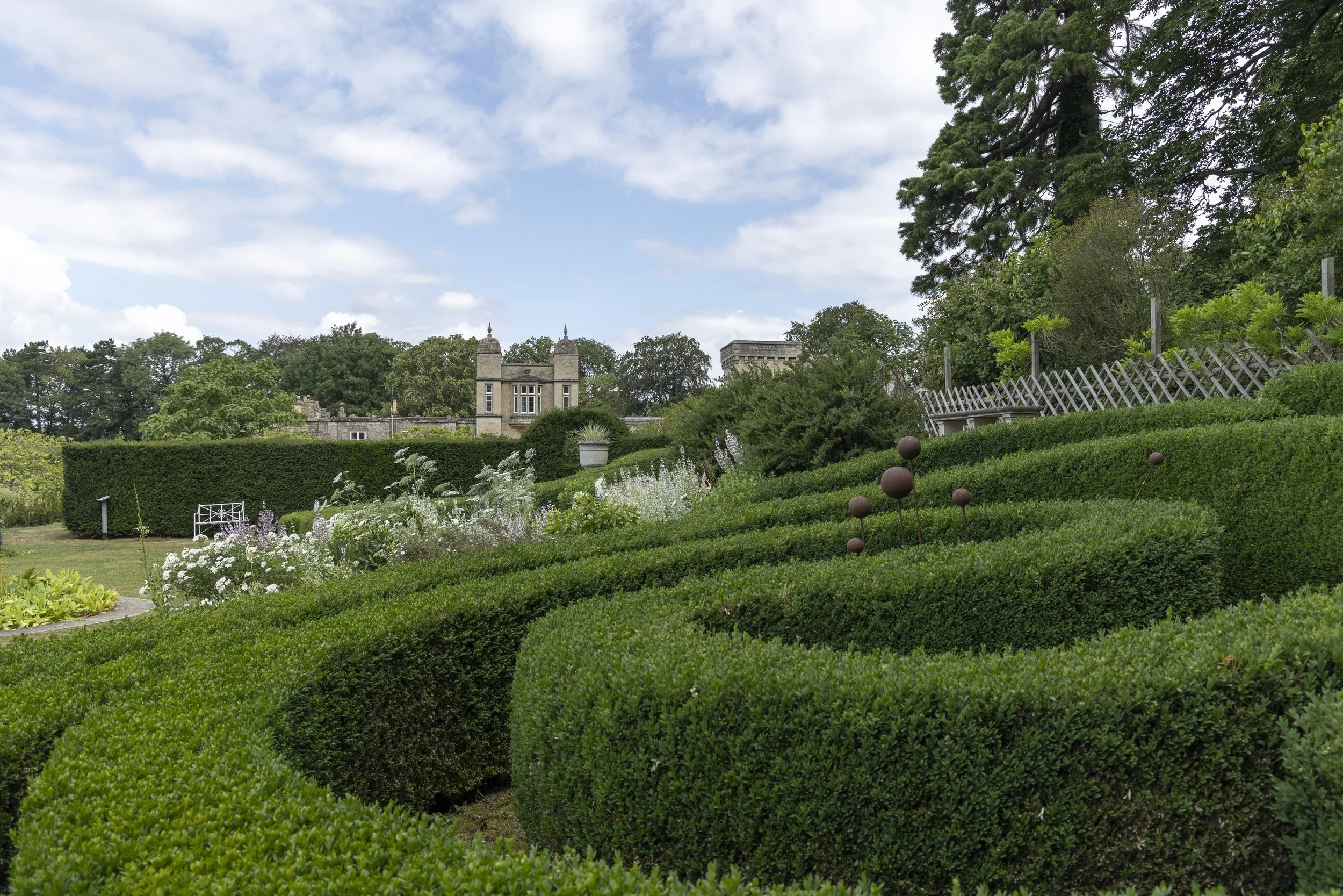 A formal garden with trimmed hedges, flowers, and decorative metal spheres, with a castle-like building and trees in the background under a partly cloudy sky.