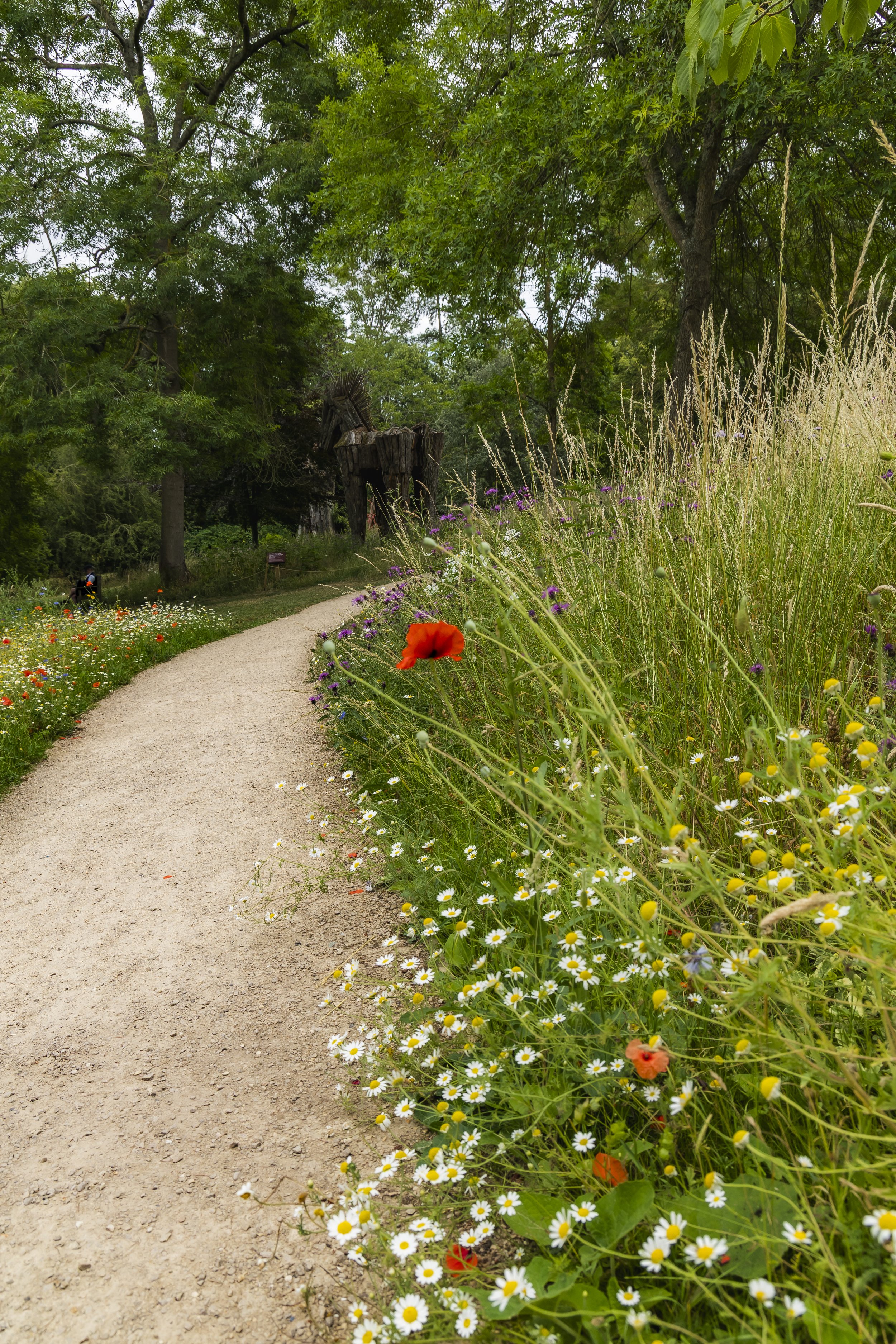 Pathway through a garden with colorful flowers on the right side, tall trees in the background, and a wooden sculpture at Burghley House.