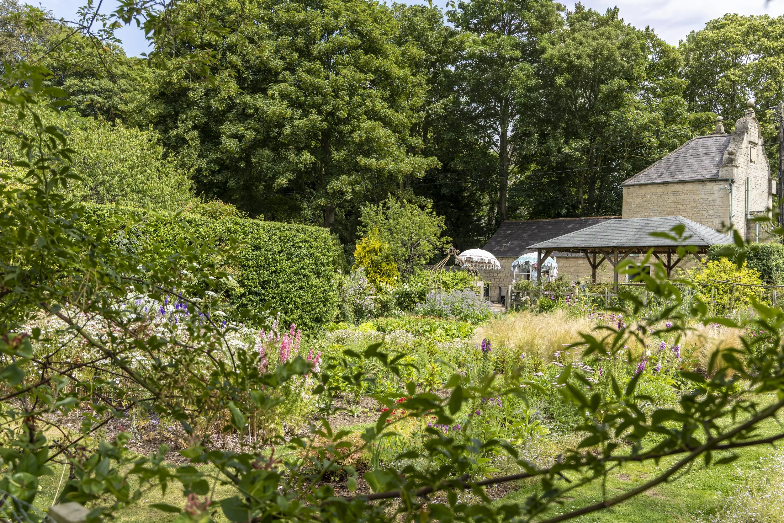 A lush garden with various flowers, shrubs, and trees, with an old stone building and a small pavilion in the background.