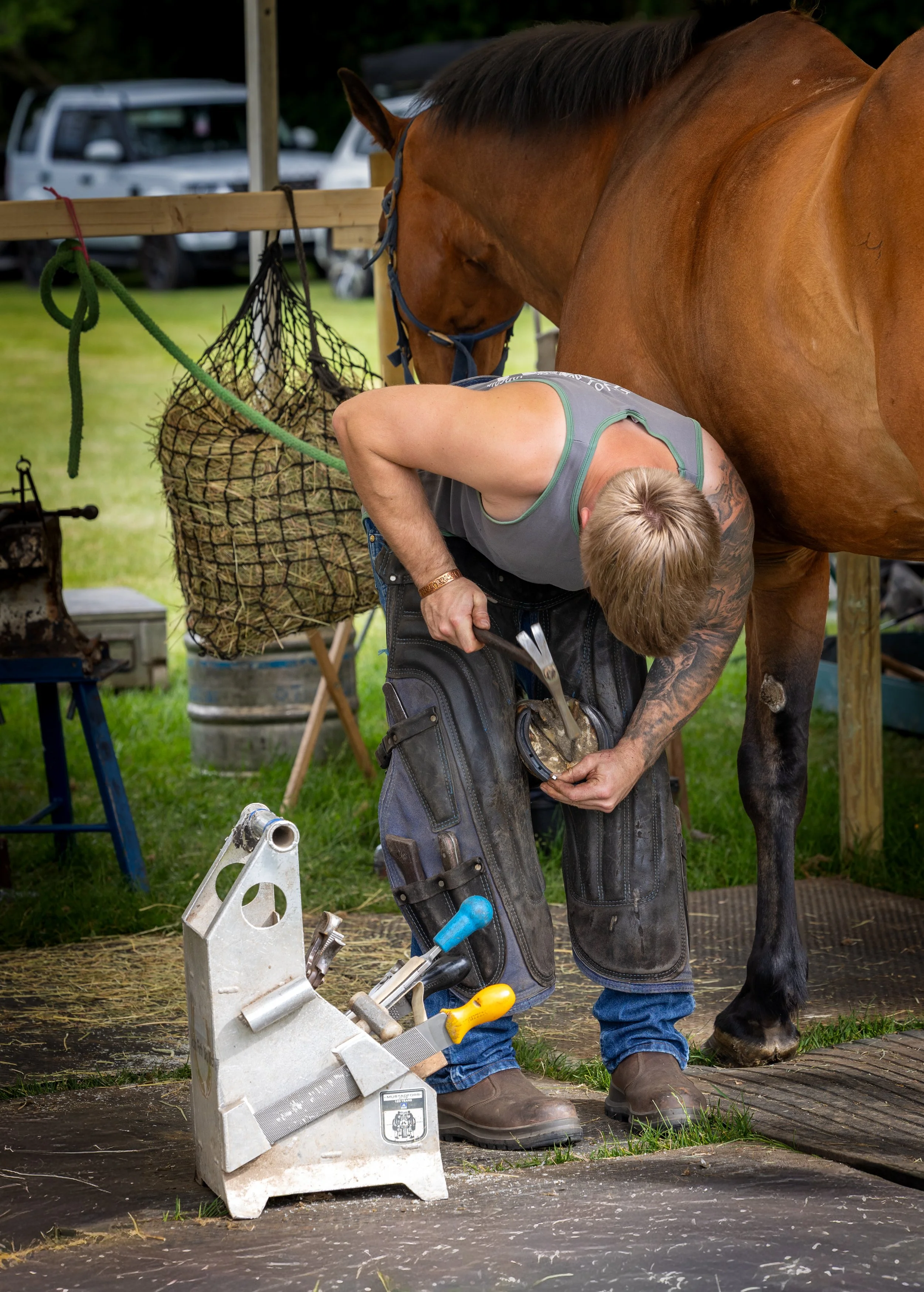 A farrier working on a horse's hoof at an outdoor farm or stable, with tools and equipment nearby.