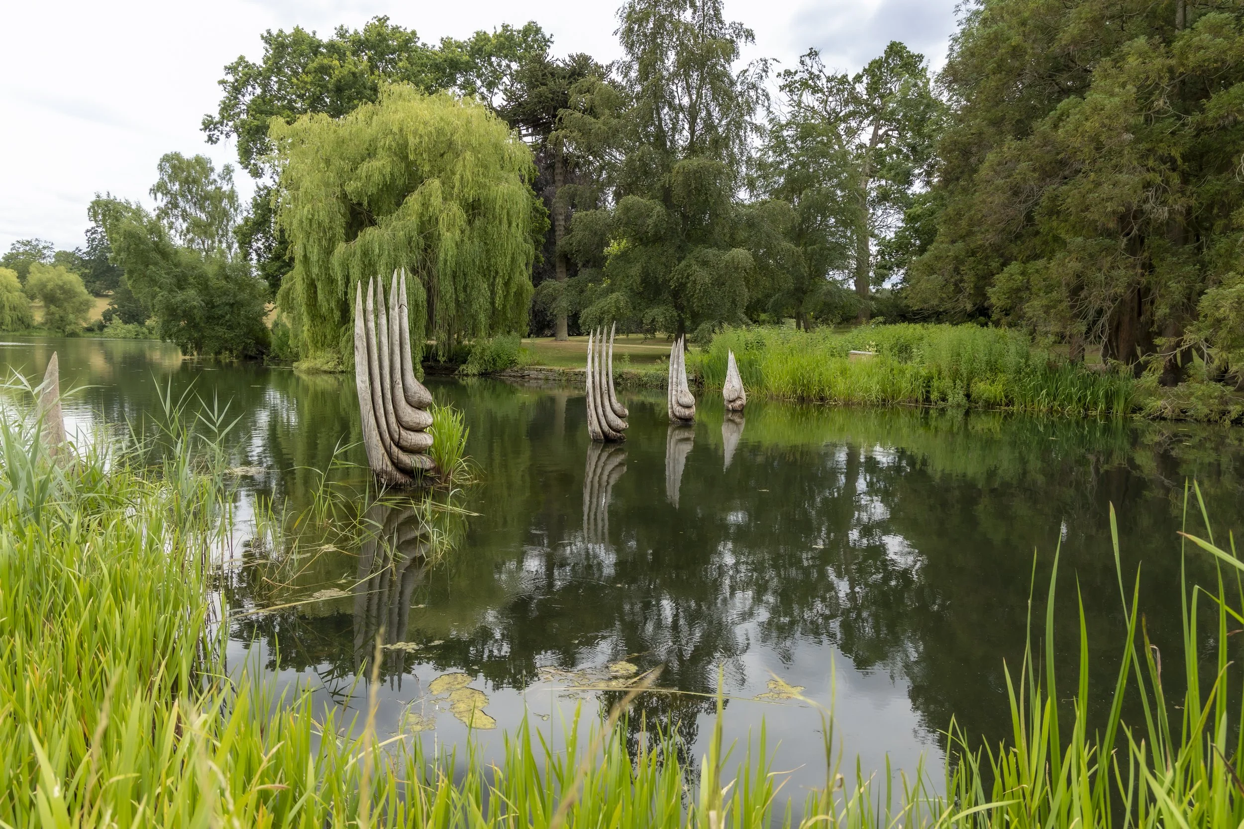 A serene lake scene surrounded by lush green trees, with wooden sculptures partially submerged in the water and dense greenery on the lakeside at Burghley House.