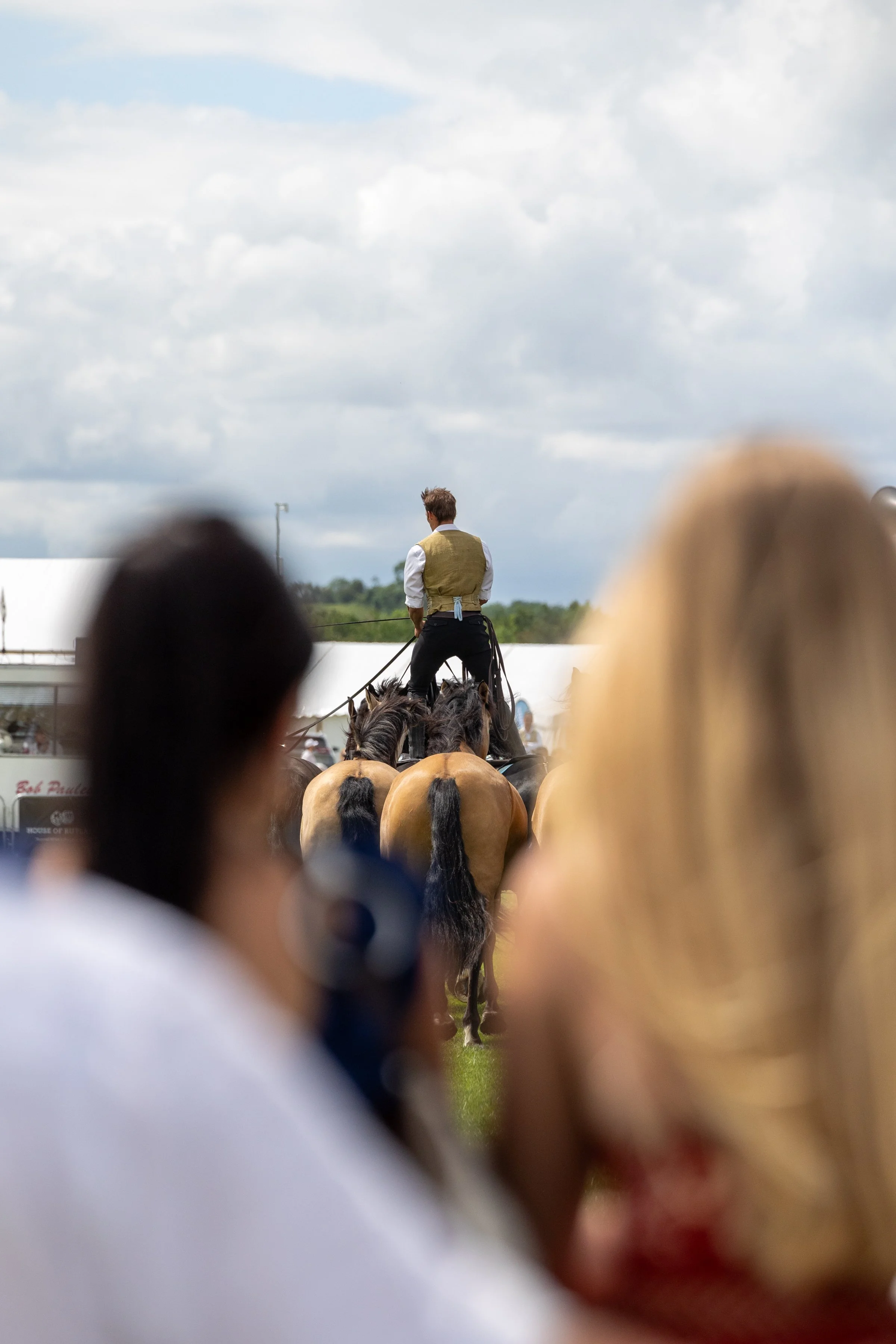 A person riding a horse with a yellow vest, seen from behind, during an outdoor event, with a cloudy sky overhead and blurred audience members in the foreground.