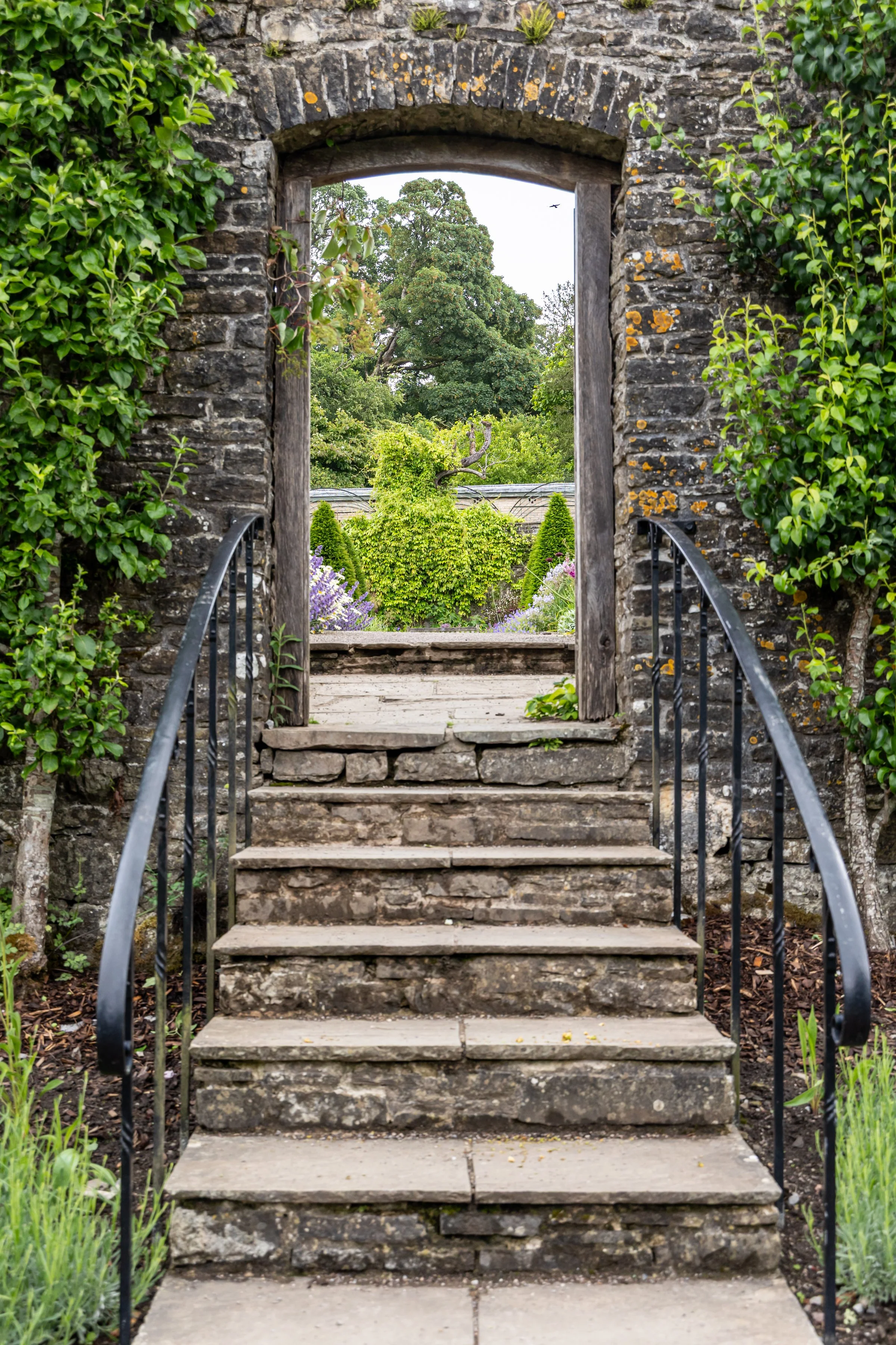 Stone steps leading up to a garden with lush green trees and plants, seen through a stone archway with a metal railing on each side.