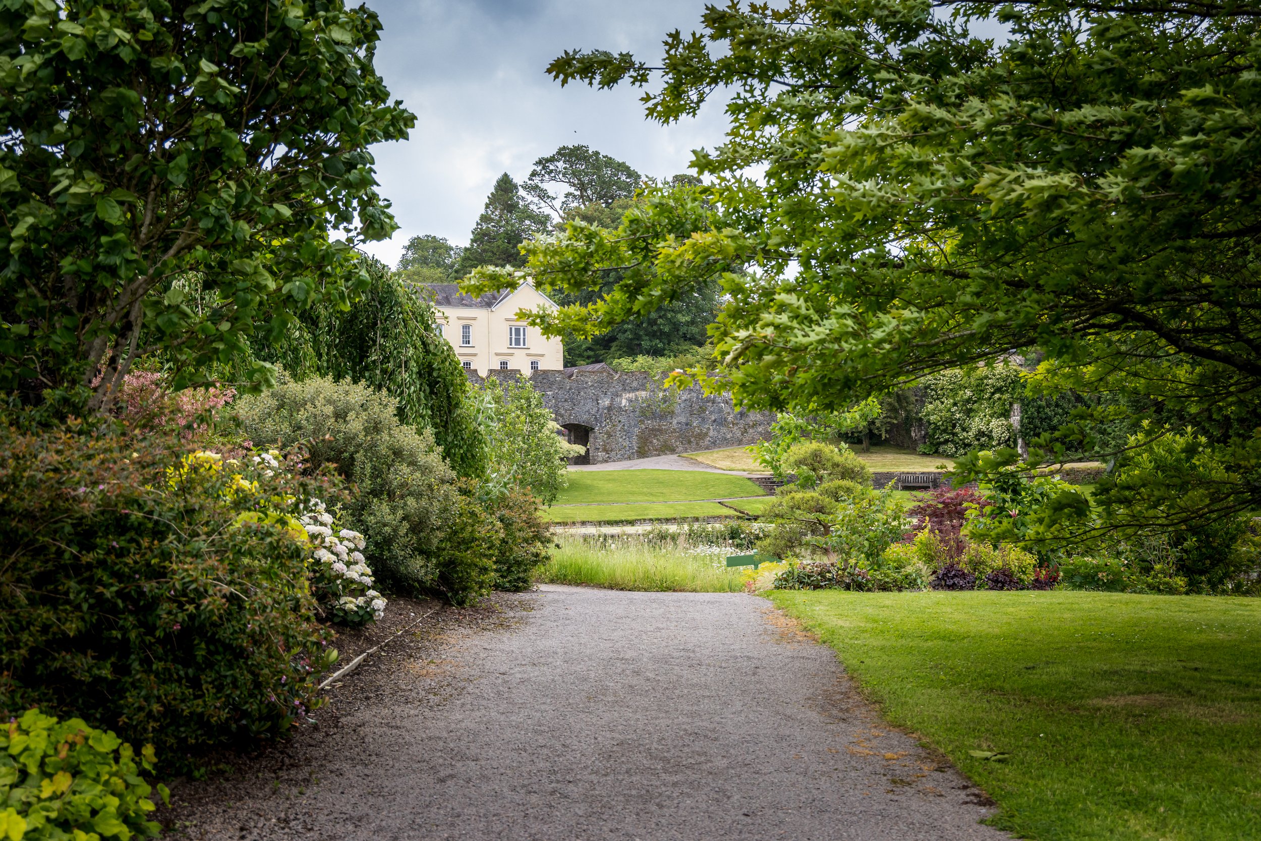 A gravel pathway winding through a lush garden with vibrant green trees and shrubs, leading to a white building and stone wall in the background.