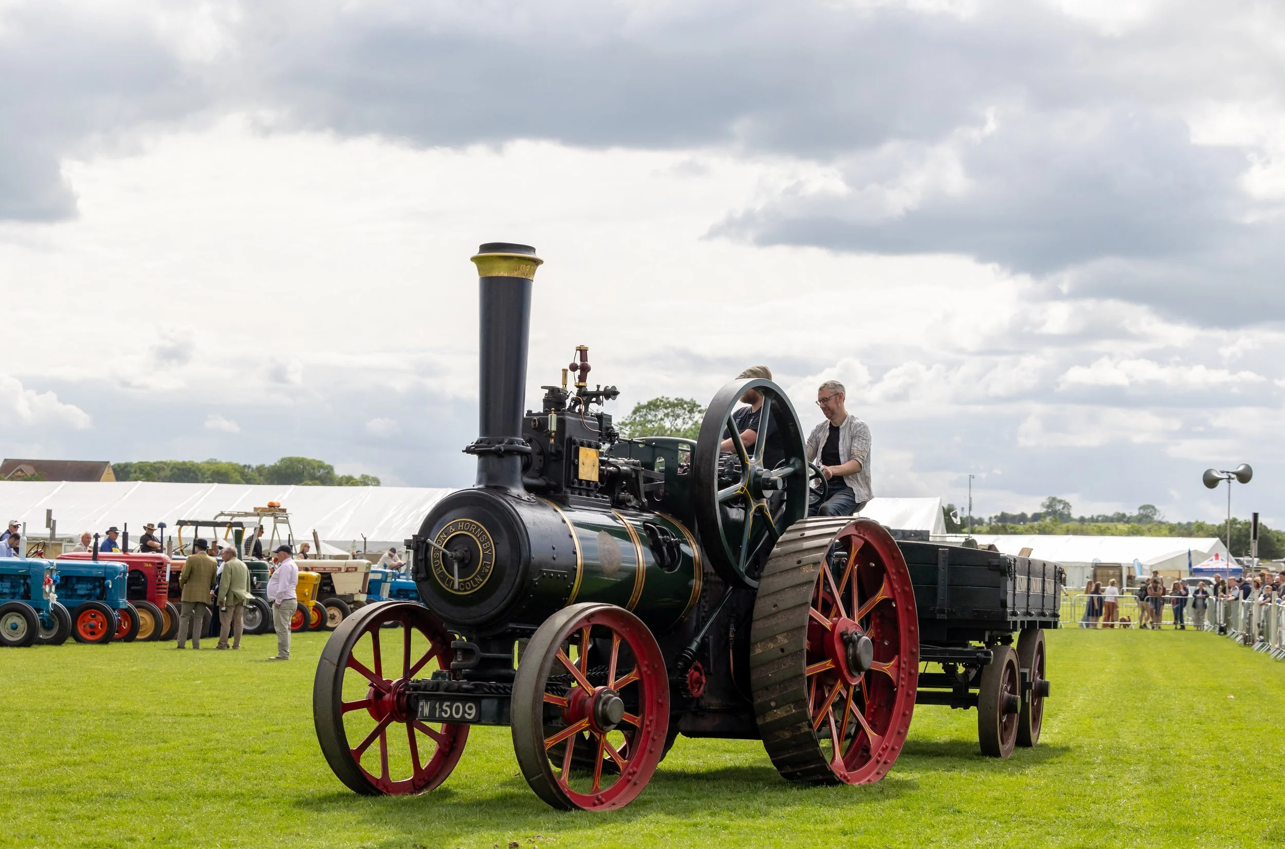 Vintage steam tractor with large red wheels displayed at an outdoor event, with people walking around and tents in the background under a cloudy sky.