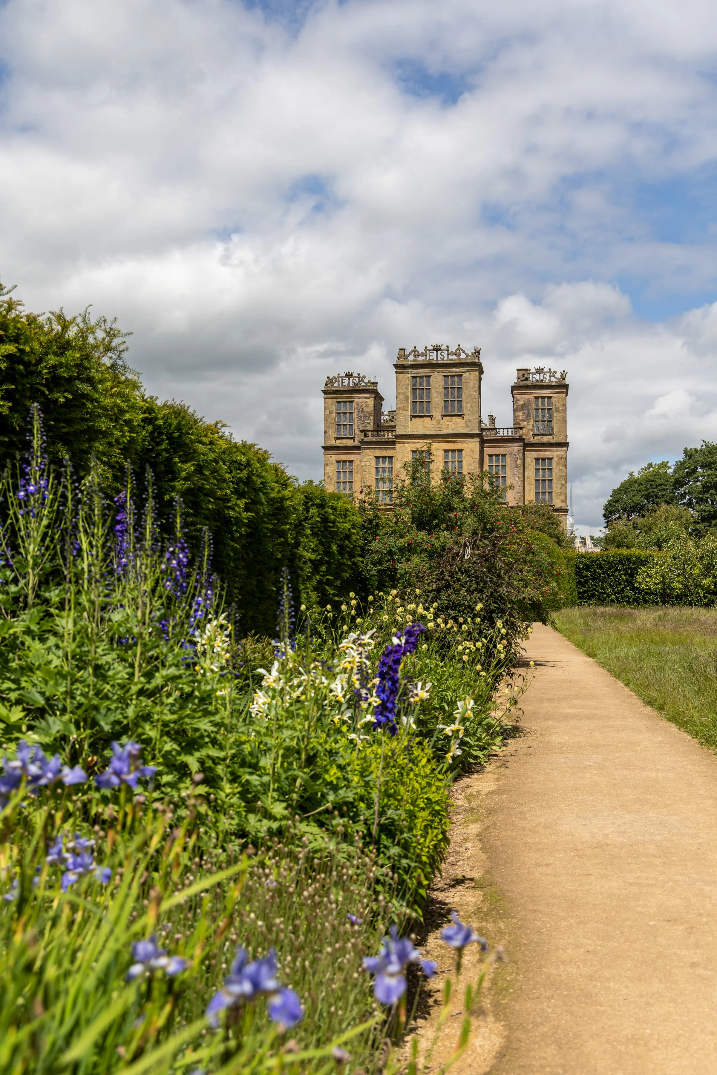 Hardwick Hall National Trust - A historic castle-like building with multiple stories and large windows, surrounded by lush green trees and shrubbery, with a clear pathway leading towards it under a partly cloudy sky.