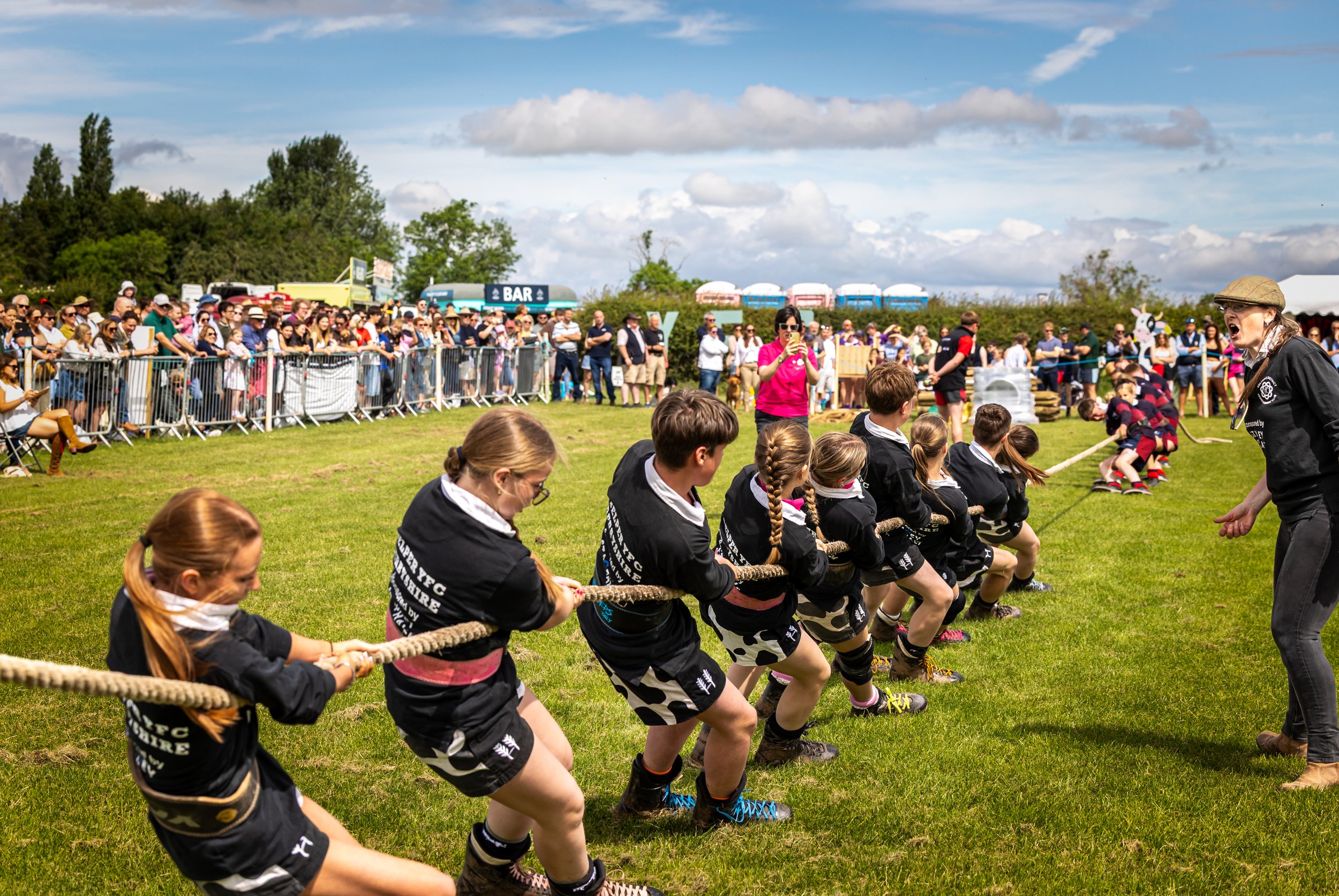 Children participating in a tug-of-war game on a grassy field during an outdoor event, with spectators watching behind a metal fence and food trucks in the background, under a partly cloudy sky.