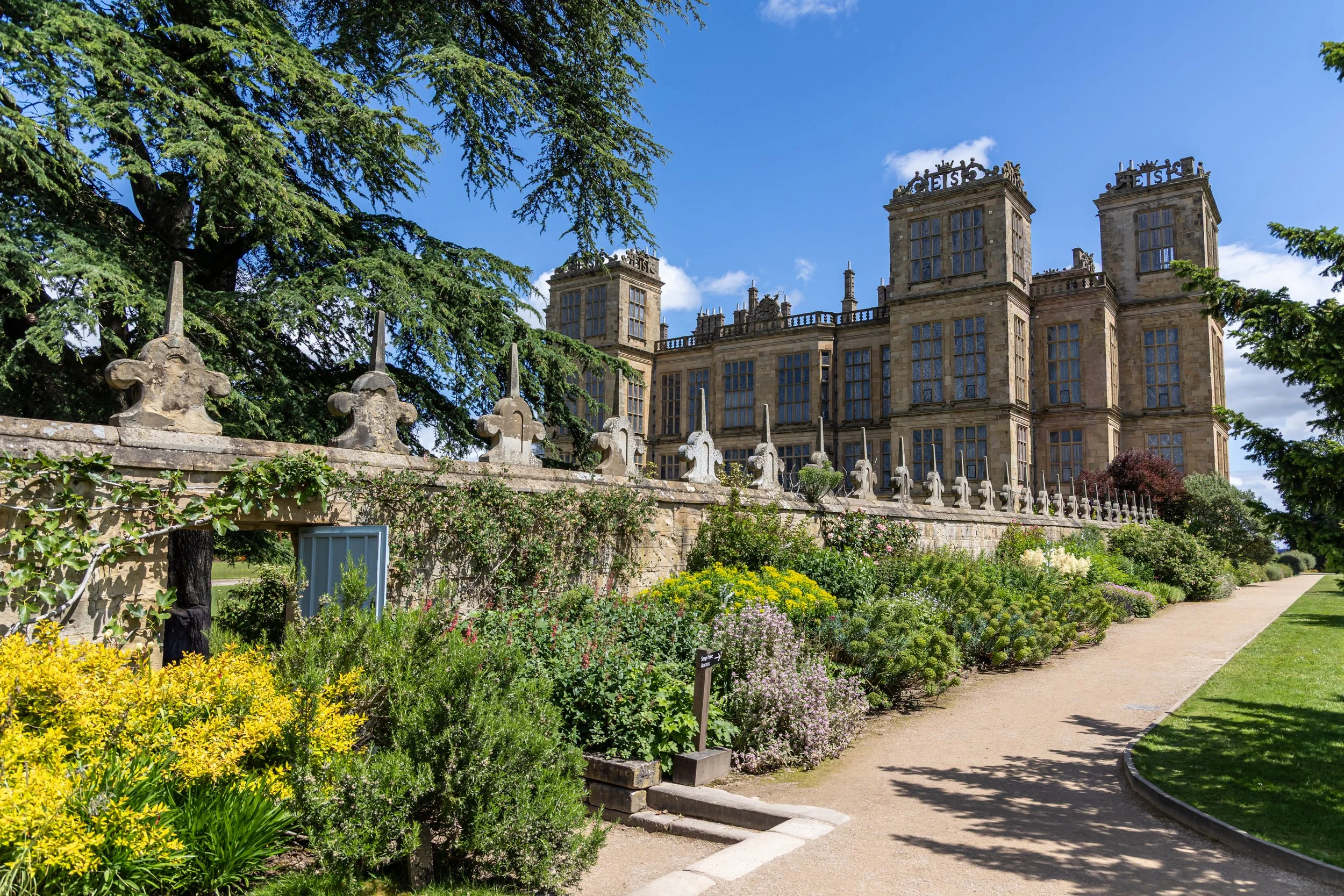 Hardwick Hall National Trust  - A historic stone building with multiple tall windows, surrounded by a lush garden with colorful flowers, trees, and a dirt pathway under a clear blue sky.