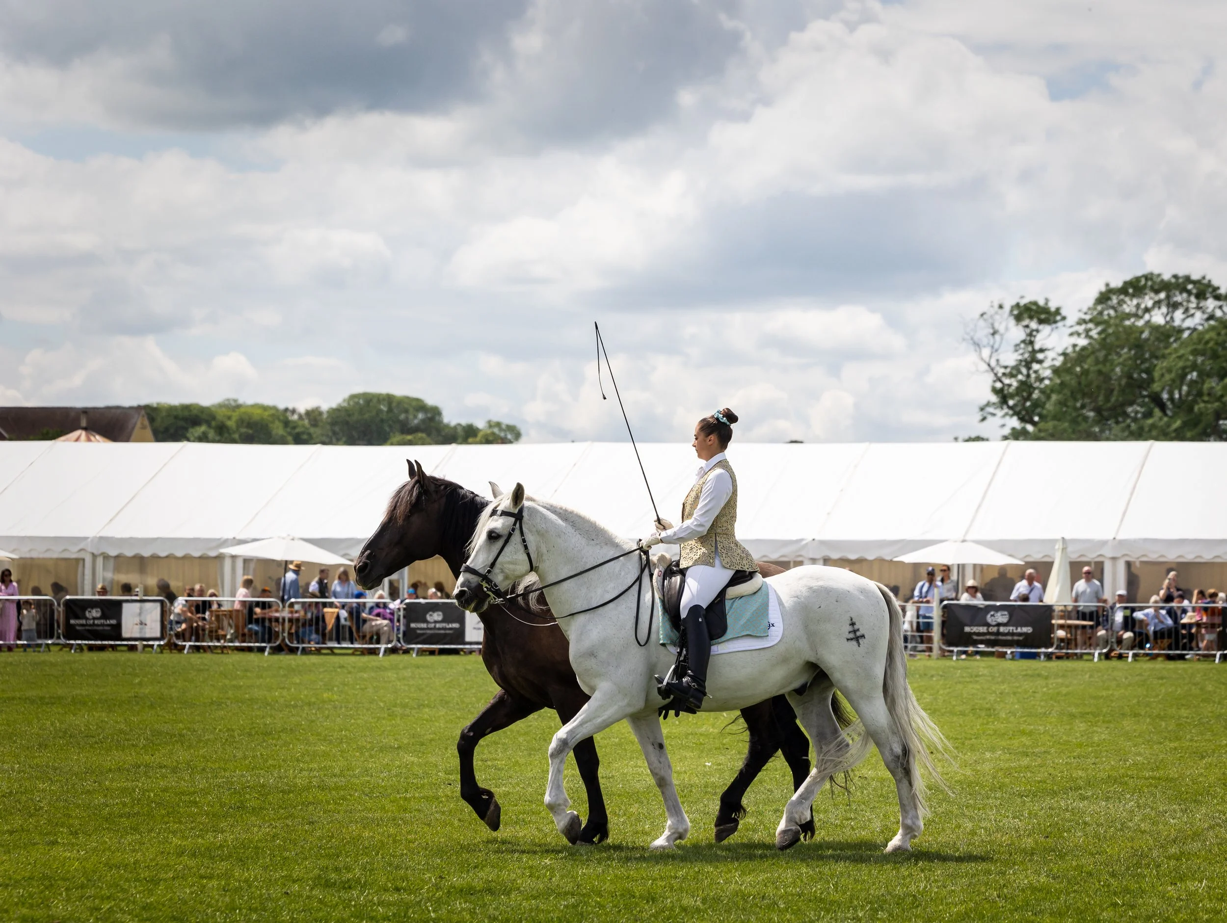 A woman in a white riding outfit and vest riding a white horse during an outdoor event under a partly cloudy sky, with a crowd watching behind a metal fence and large white tents in the background.