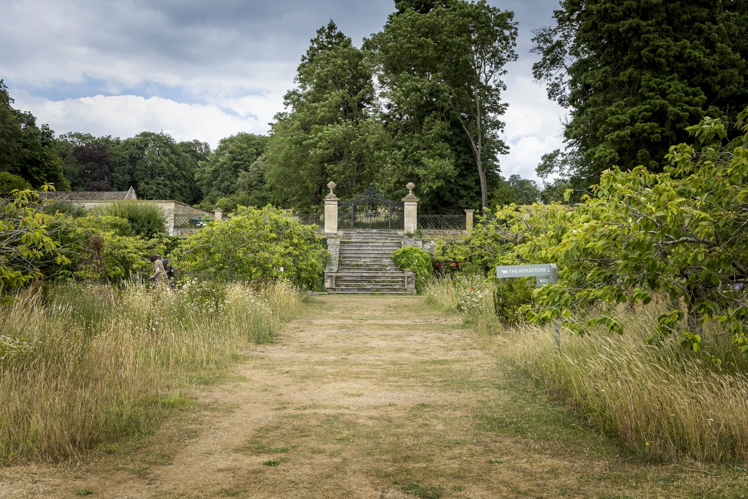 A garden or park scene with a dirt pathway leading to a stone staircase and a black wrought-iron gate, surrounded by lush greenery, trees, and a cloudy sky.
