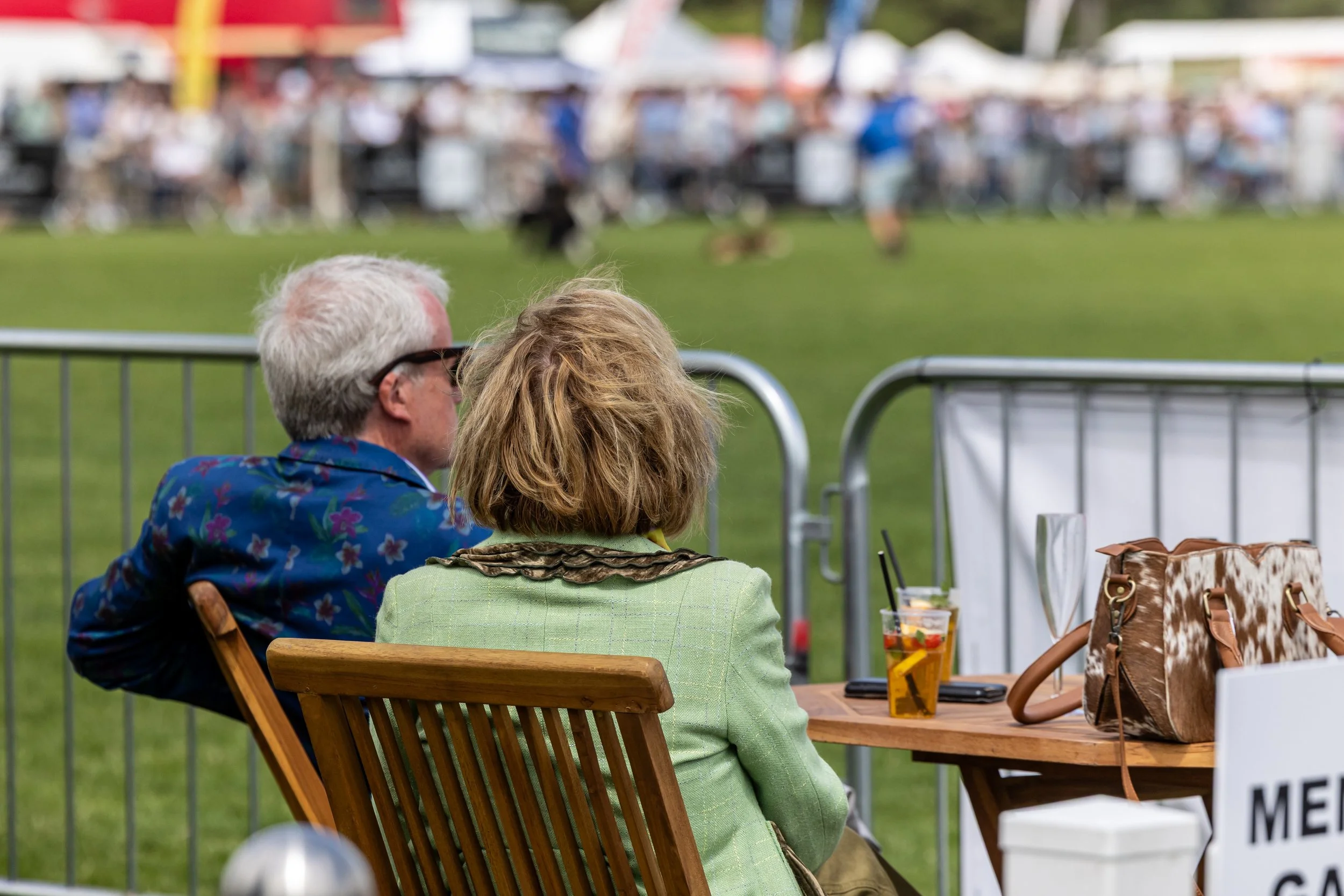 Two women sitting at an outdoor table, viewed from behind, watching a fair or carnival, with blurred crowds and tents in the background.