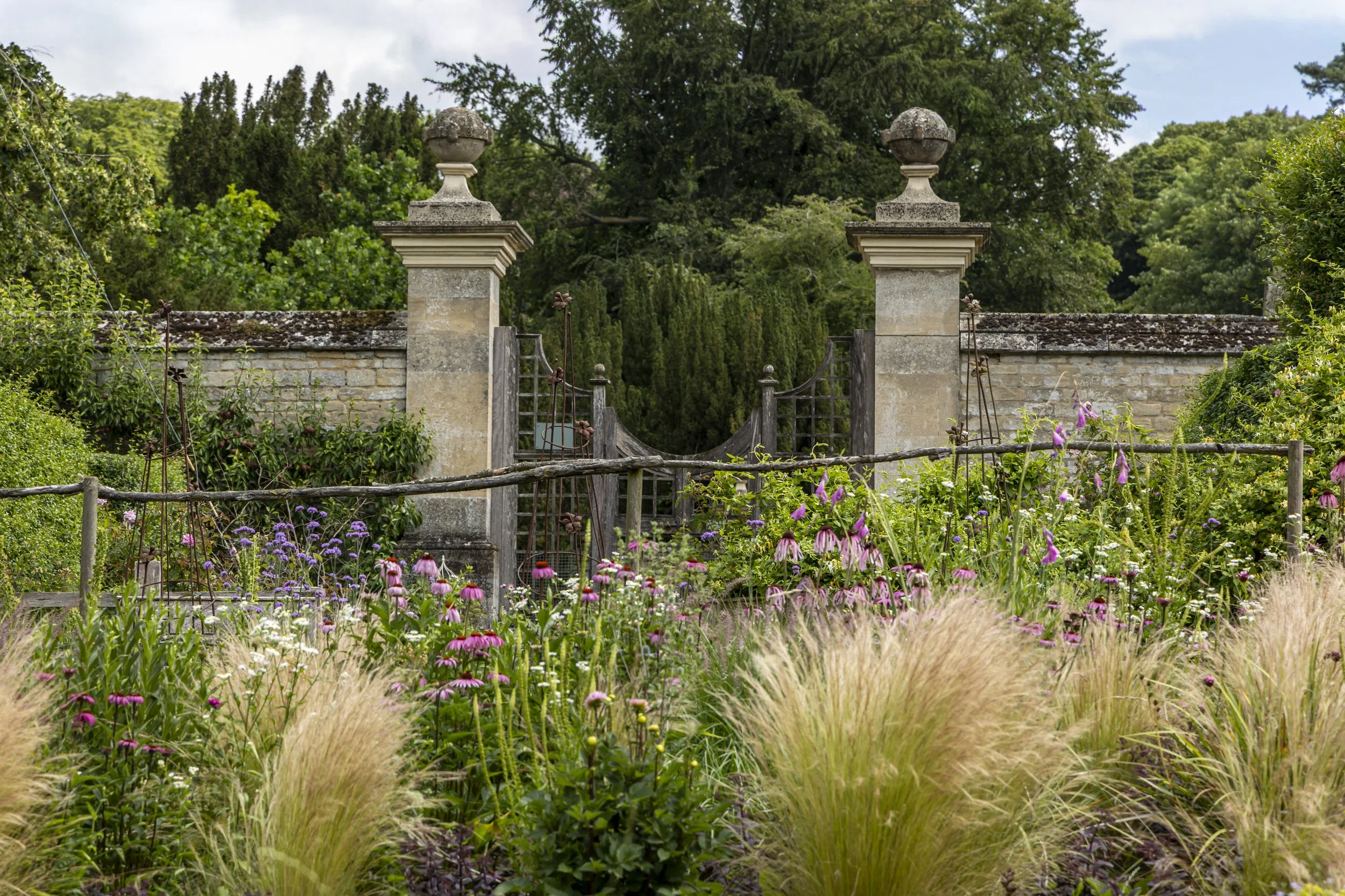 Colored flower garden in front of a stone wall with two stone pillars and gates, surrounded by lush green trees and bushes.
