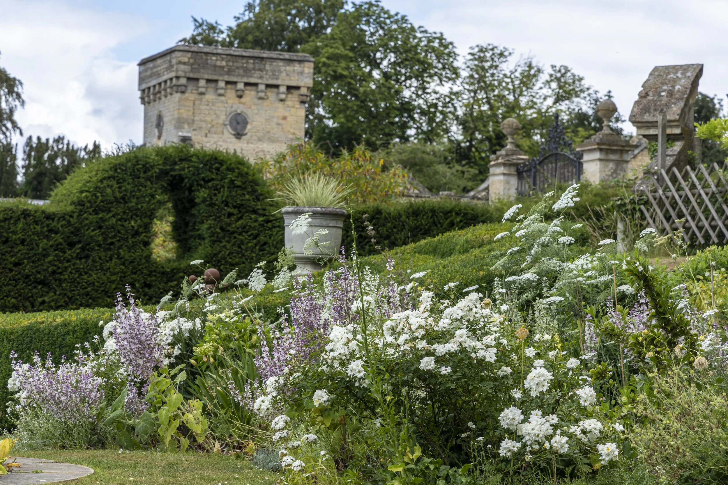 A lush garden with various white and purple flowers in the foreground, green bushes, and a stone tower with a clock in the background. There are also some ornate stone features and wooden fencing.