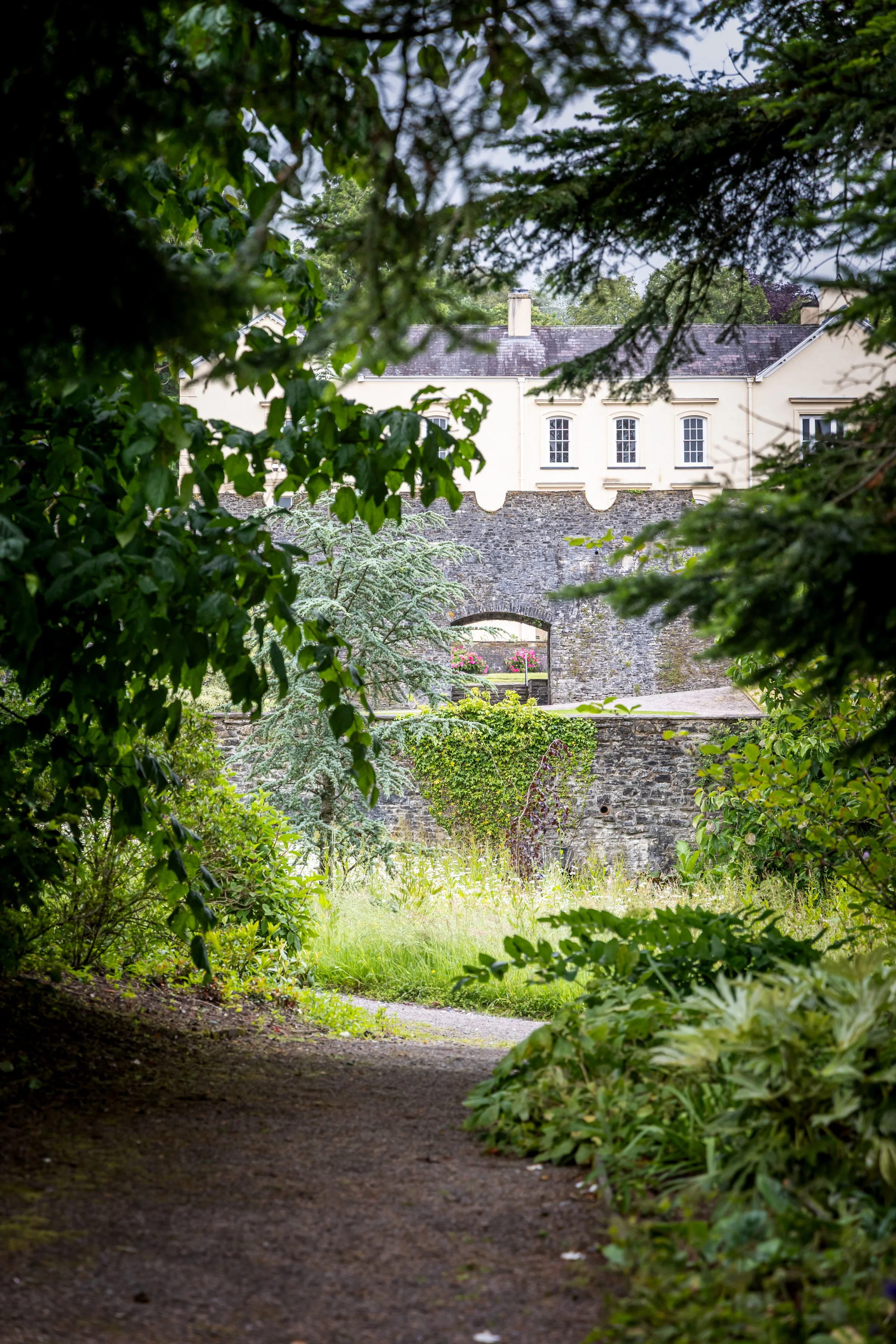 A garden pathway leading towards a stone bridge with a cream-colored house with large windows and a gray roof in the background, surrounded by trees and greenery.