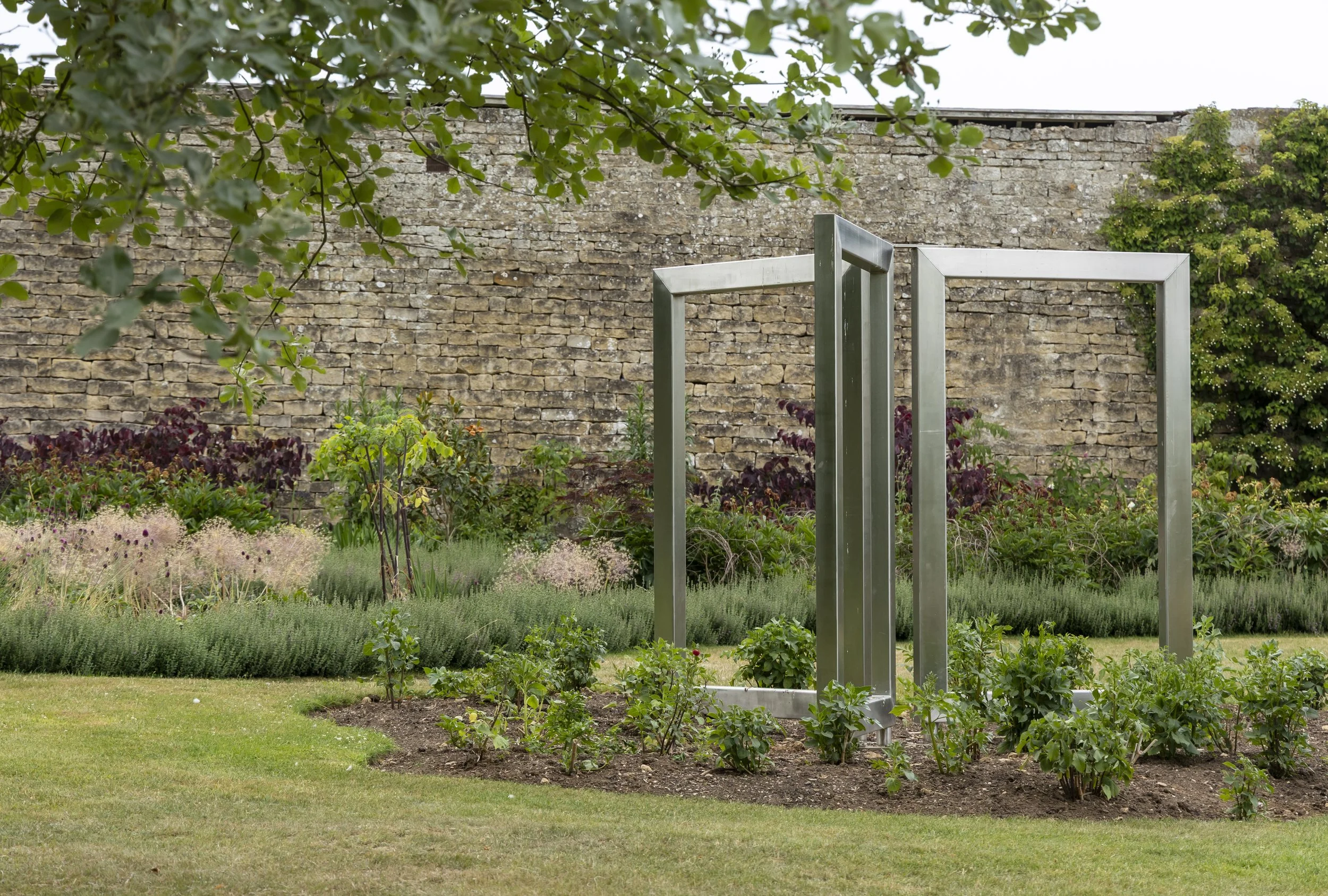 A garden with various plants and shrubs in front of a brick wall, featuring a modern metal garden structure.