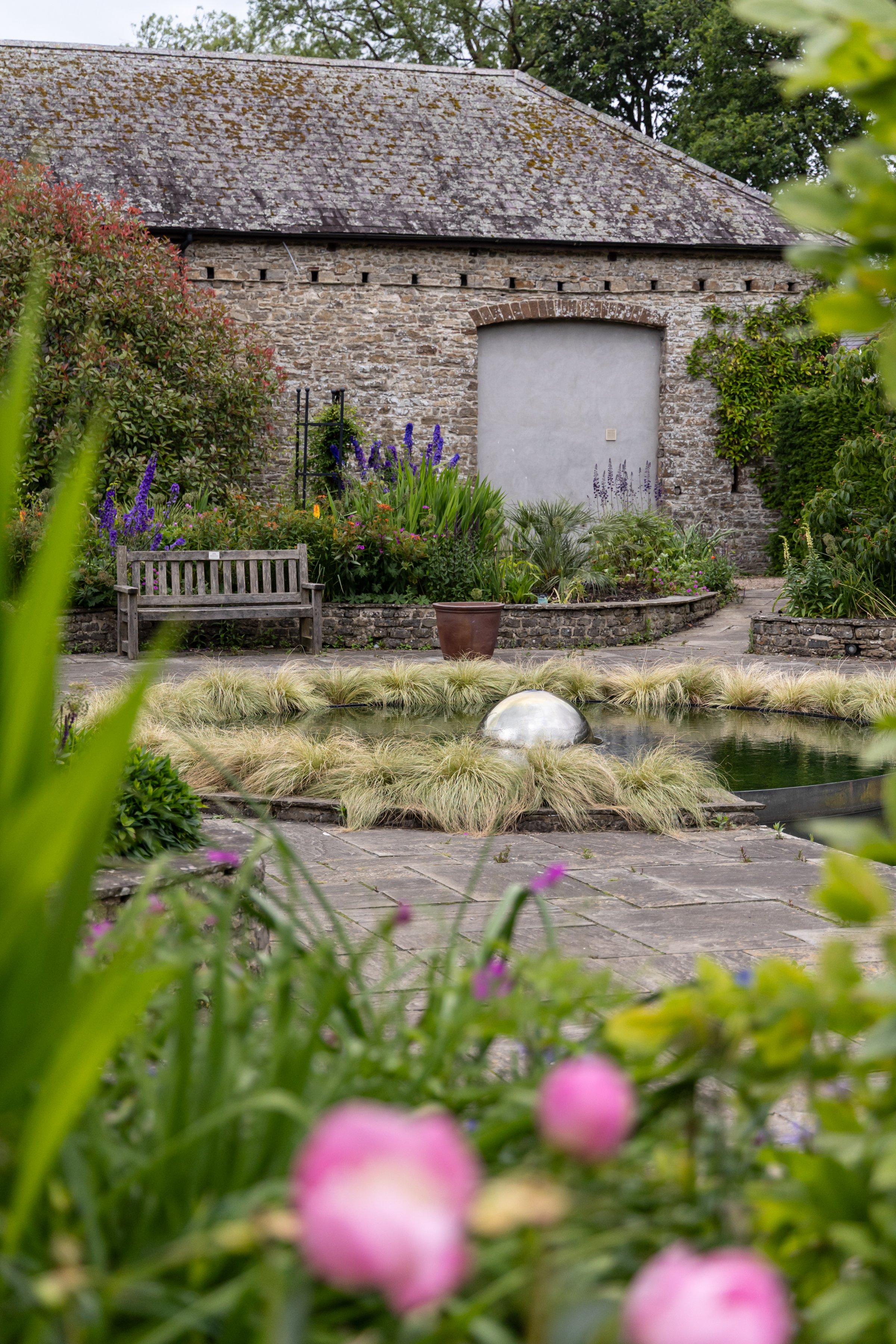 A garden scene with a pond surrounded by plants and flowers, a wooden bench, and a stone building with a pitched roof in the background.