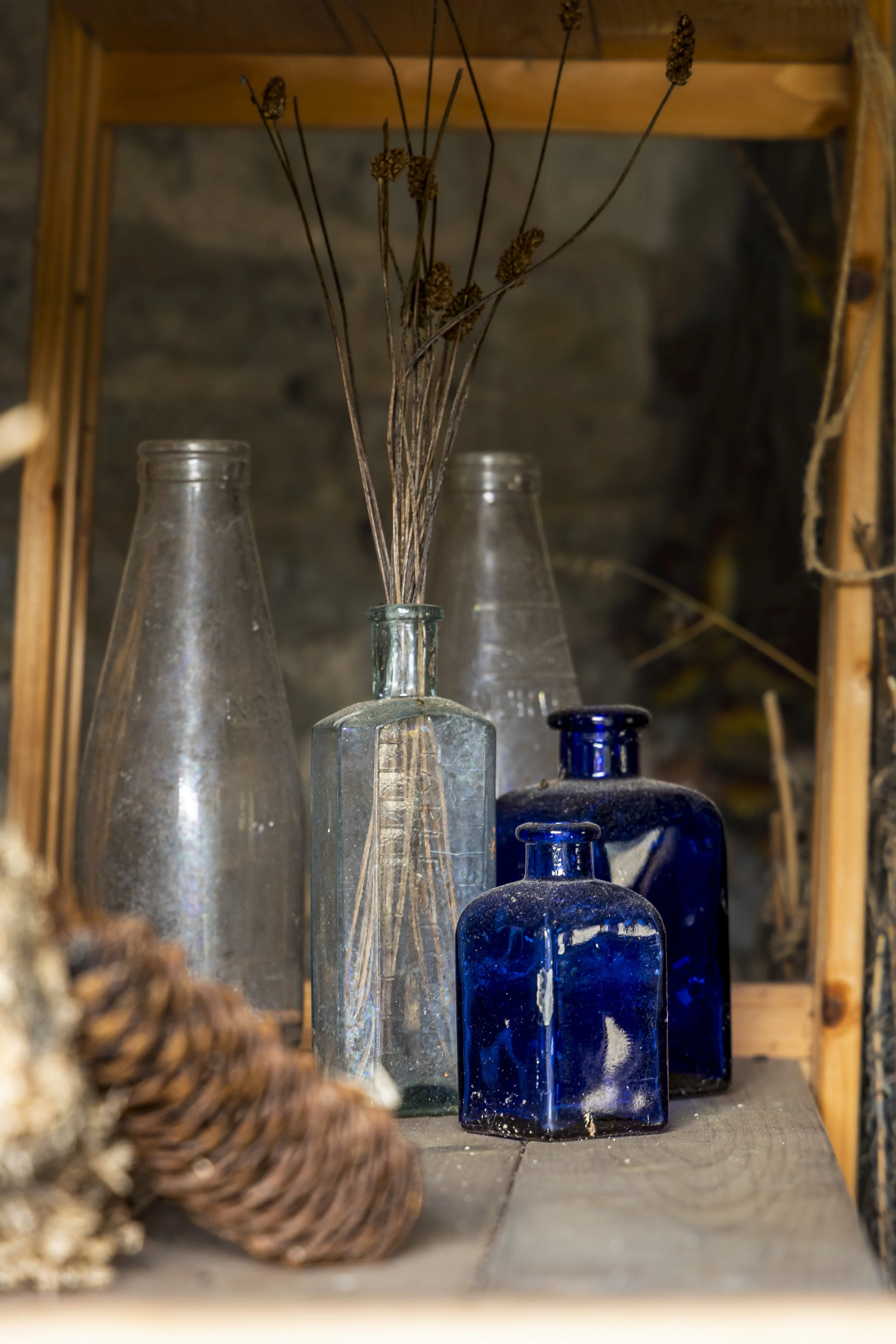 A collection of vintage glass bottles, including clear and cobalt blue, arranged inside a wooden shelf unit with a rustic background, and dried plant stems placed in one of the bottles.