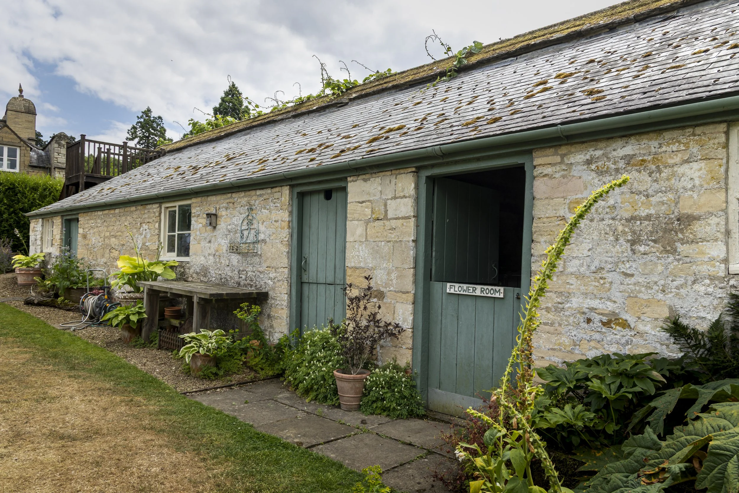 A stone cottage with a sloped slate roof partially covered in moss and vine plants. The front has a door and a window labeled 'Flower Room', surrounded by potted plants and garden greenery.