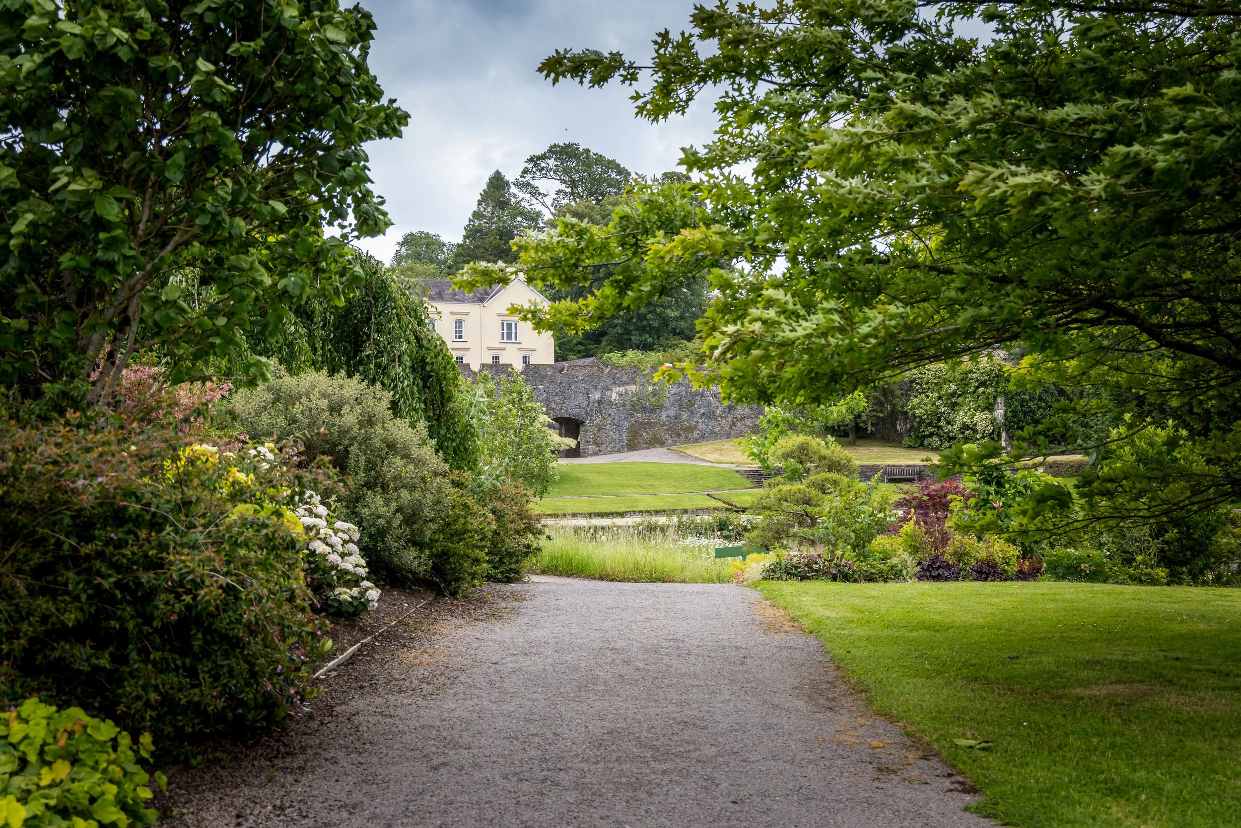 A garden pathway surrounded by lush green foliage and blooming flowers, leading to a stone wall and white house in the background under a partly cloudy sky.
