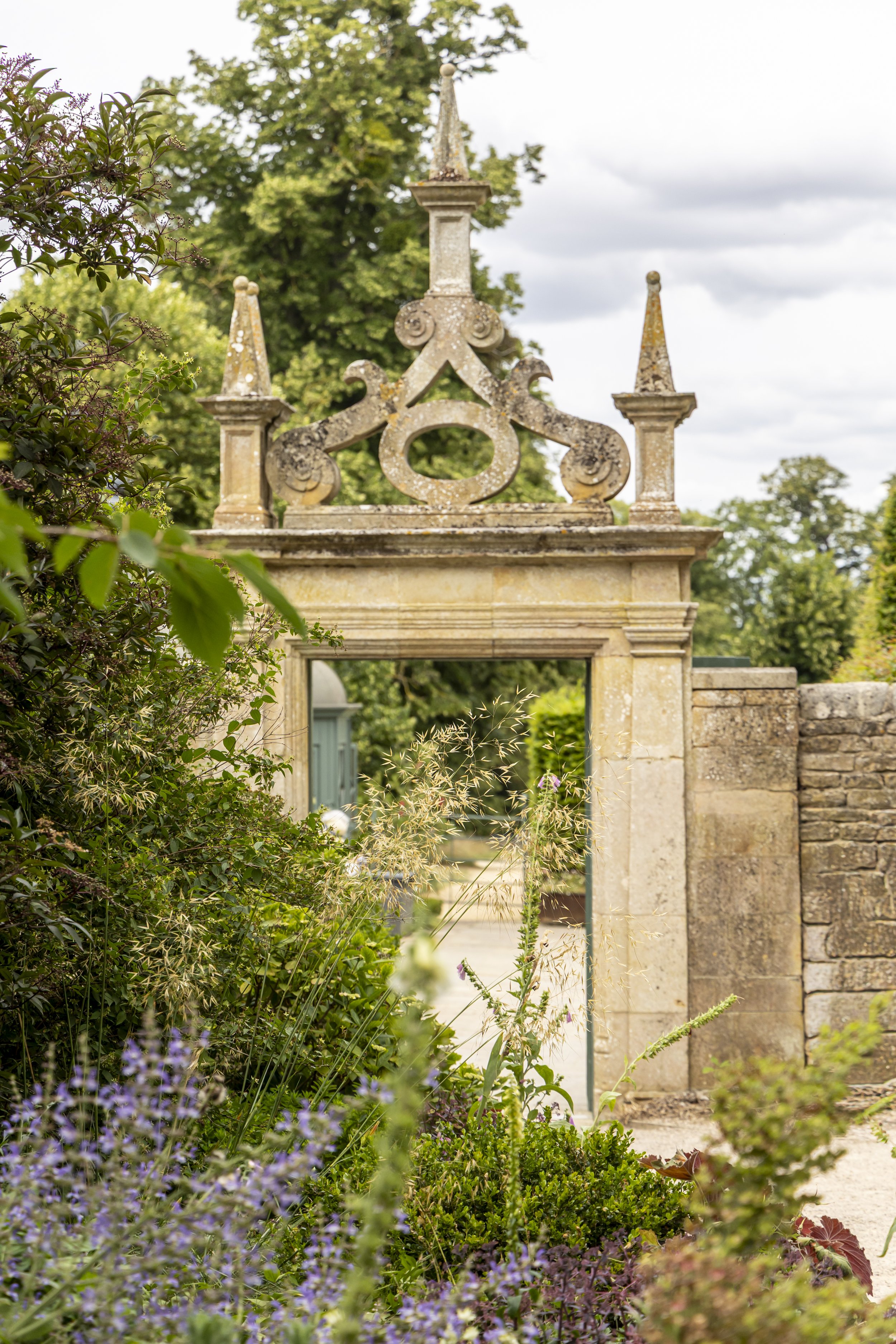 Stone gate with decorative top and three spires, surrounded by lush green plants and flowers, with trees and cloudy sky in the background at Burghley House.