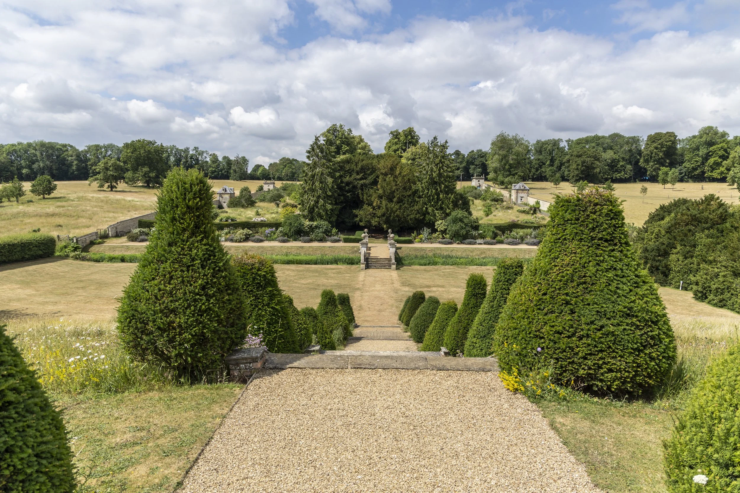 View of a formal garden with trimmed bushes, stone steps, and a pathway leading to open fields and trees under a partly cloudy sky.