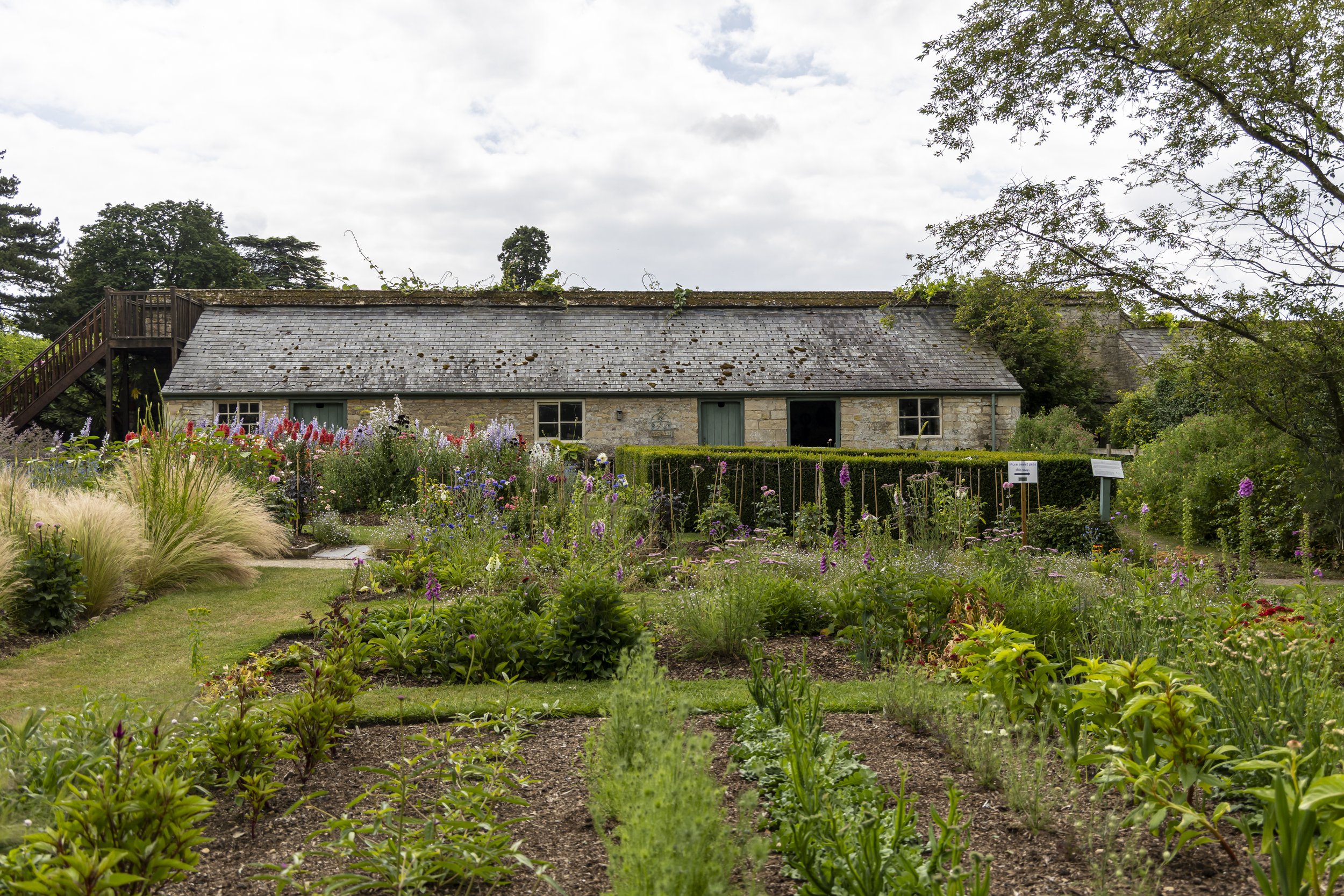 A large stone cottage with a slate roof surrounded by colorful flower gardens and green trees.