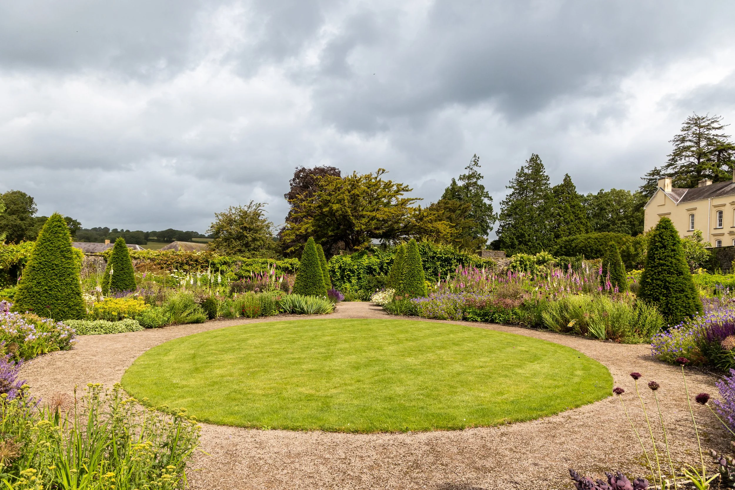 A lush garden with a circular lawn, colorful flower beds, and a gravel pathway. There are green trees, a white house, and a cloudy sky in the background.