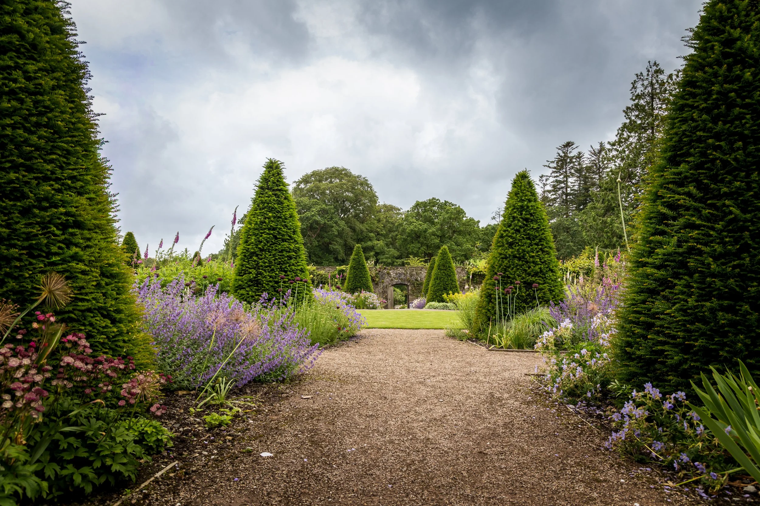 A garden pathway lined with colorful flowers and tall, cone-shaped evergreen trees, leading to an archway and lush green trees under a cloudy sky.